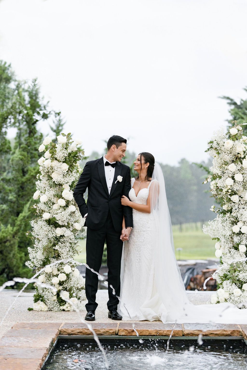 Newlyweds pose together on a platform with cascading white floral arrangements and a water feature.