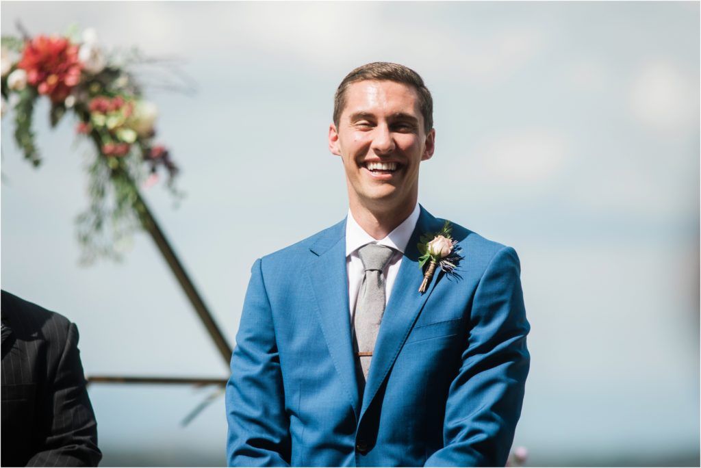 Groom in bright blue suit smiles during outdoor wedding ceremony with floral arch.
