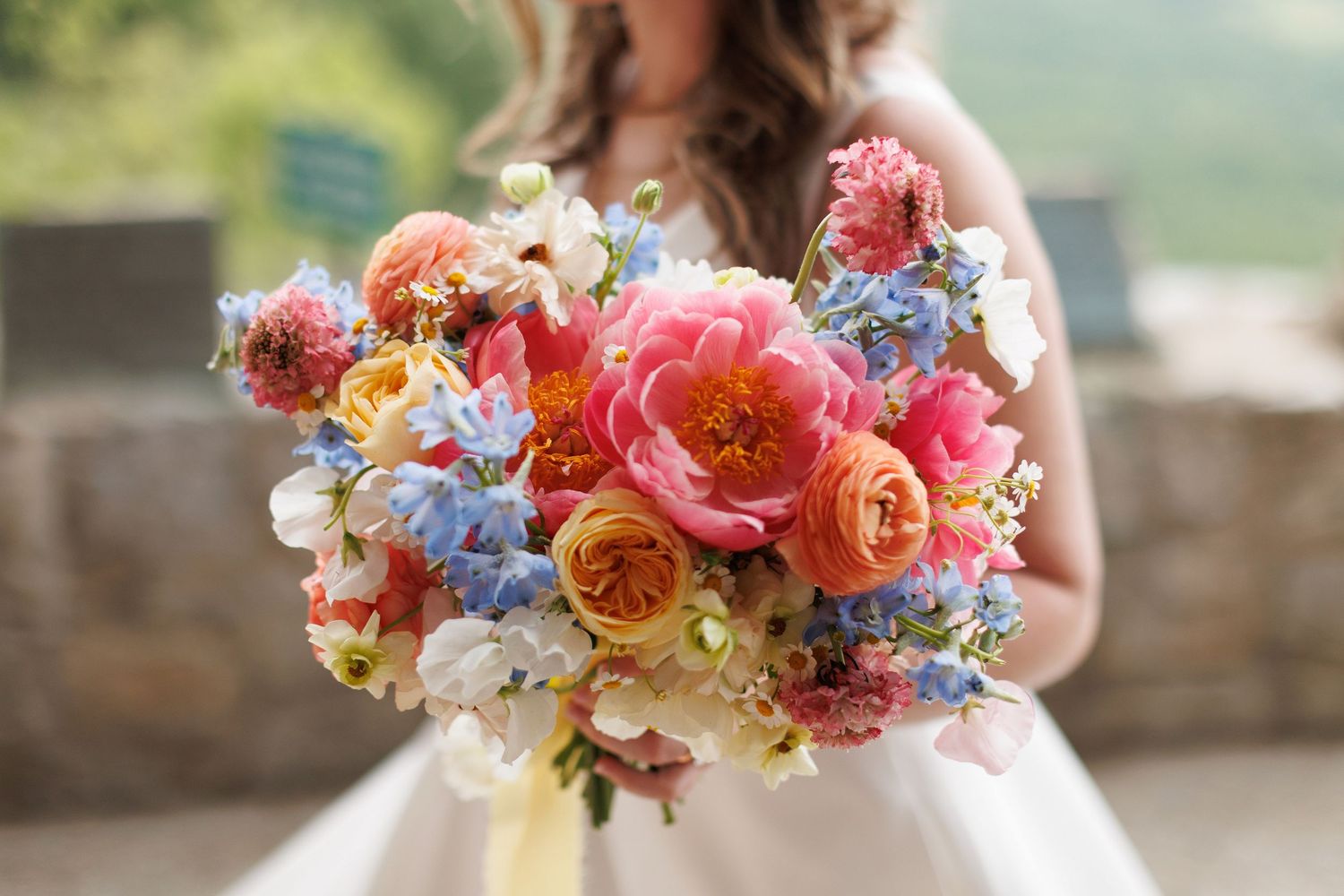 A vibrant bridal bouquet featuring coral peonies, orange roses, blue delphinium, and white florals held against a white dress.