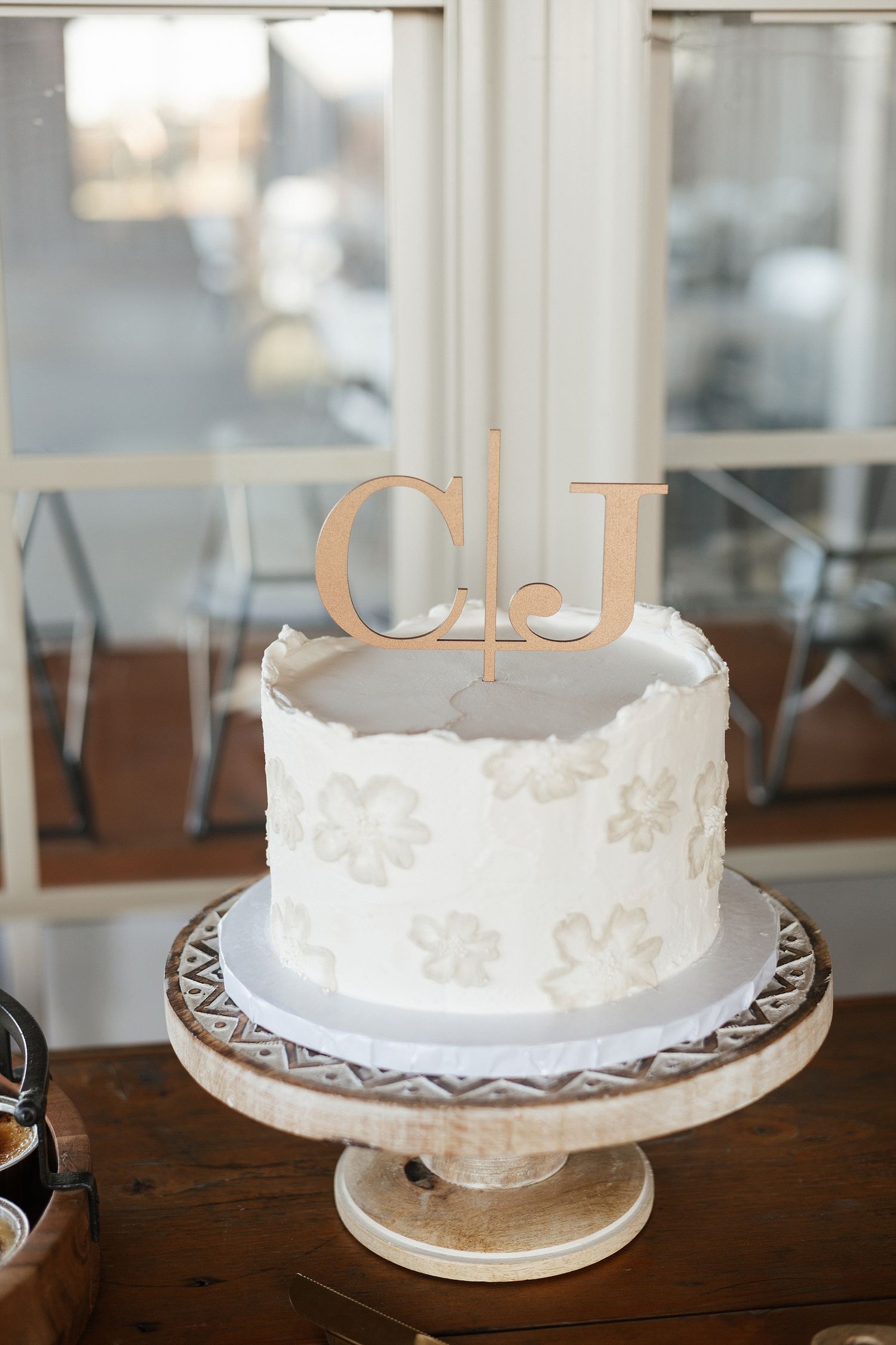 Elegant white wedding cake with delicate lace pattern design and gold monogram 'CJ' topper on a decorative cake stand.