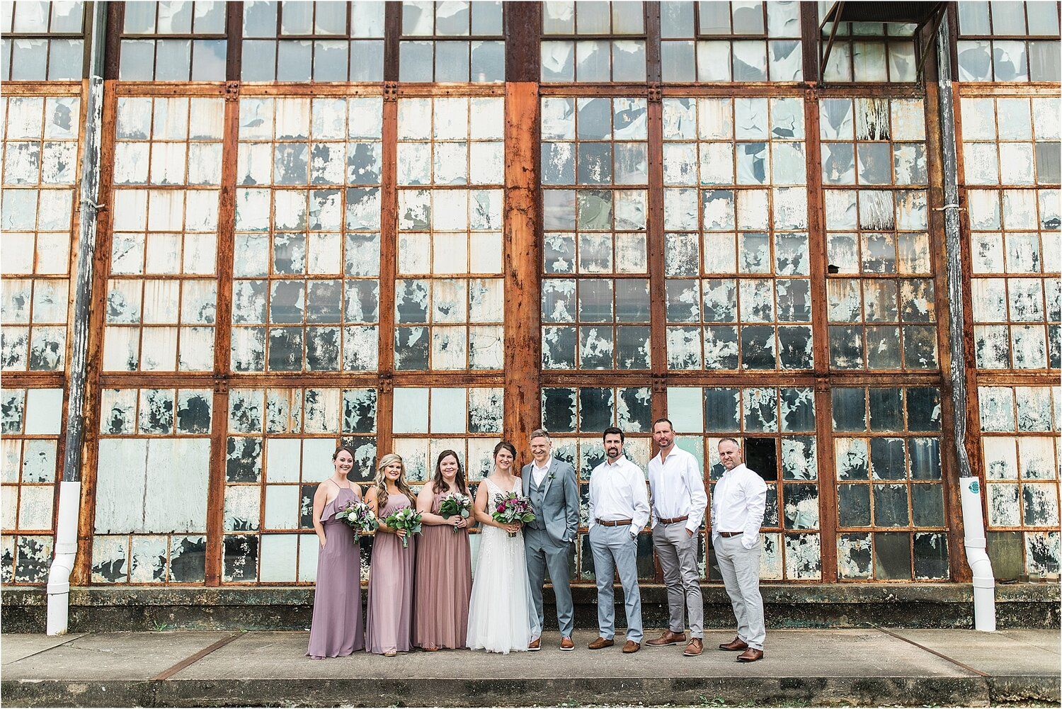 Wedding party poses in front of industrial windows at rustic venue.