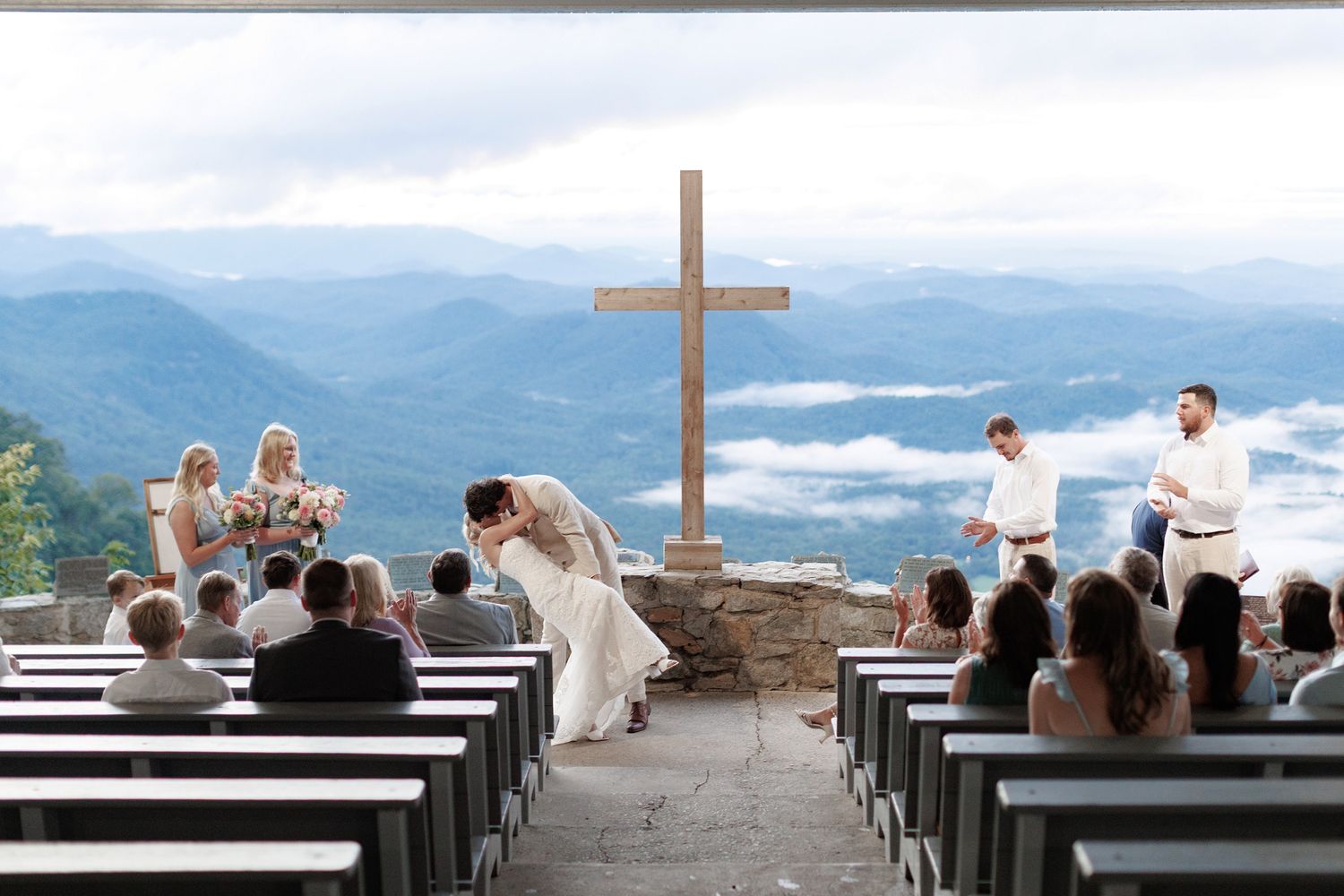 Wedding guests seated on benches observe an outdoor mountaintop ceremony with a large cross and mountain vista.