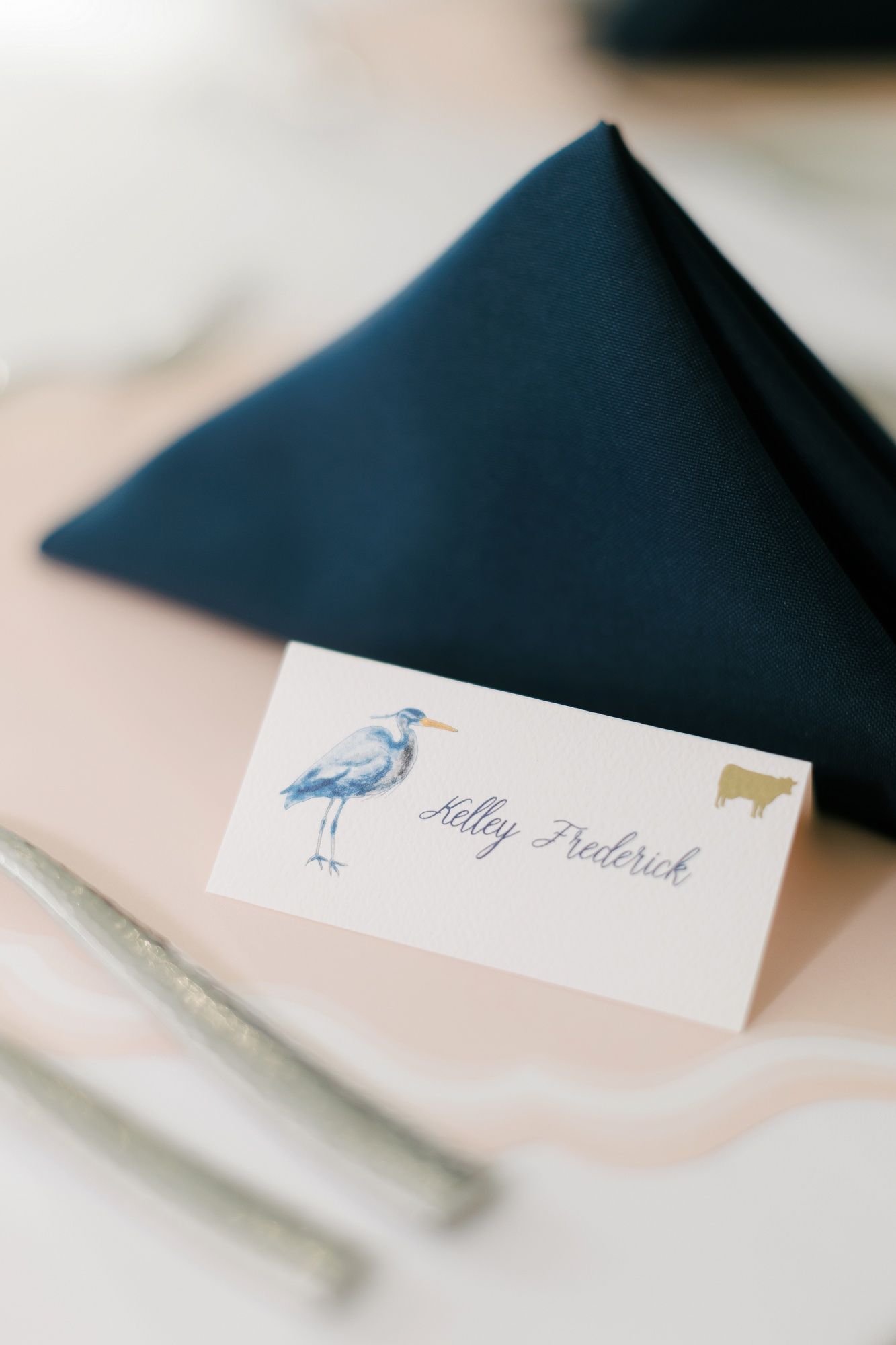 A navy blue wedding place card features a delicate blue heron illustration and elegant calligraphy.