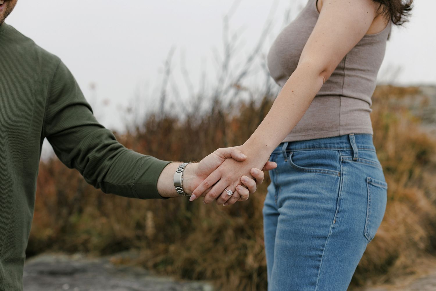 Two people holding hands while wearing casual clothing, showing an engagement ring outdoors.