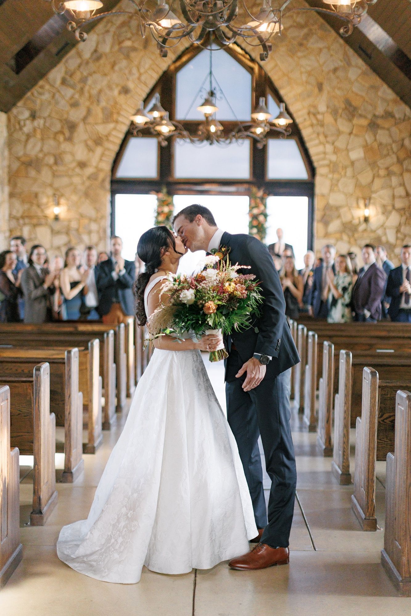 Couple shares romantic moments in a rustic stone chapel with wooden pews and elegant chandeliers.