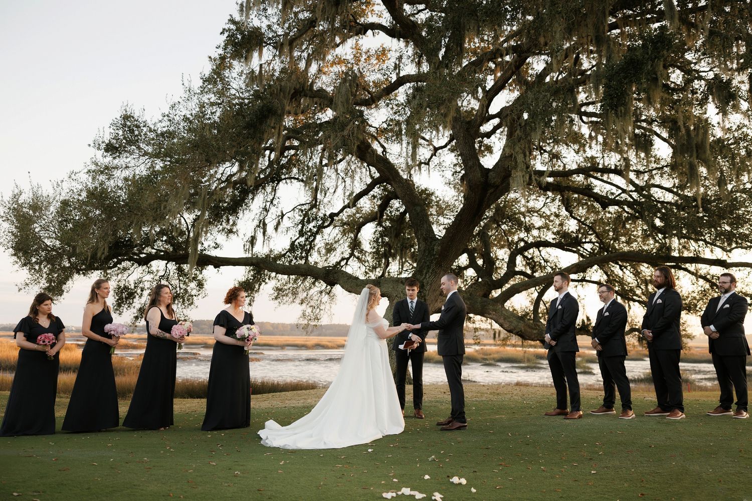 Wedding party arranged in line under sprawling oak trees at sunset.