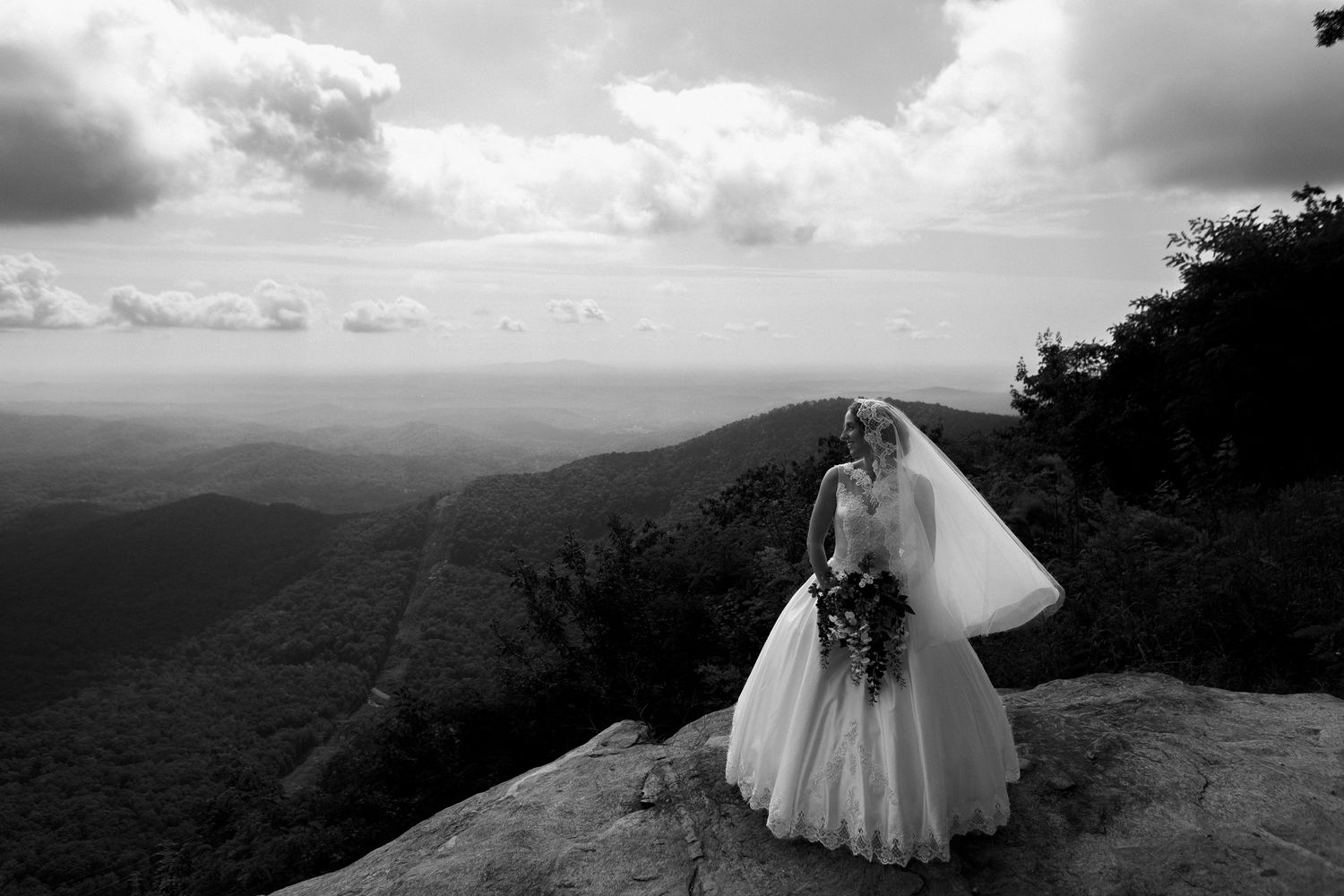 Black and white photo of bride in ball gown standing on rocky mountain overlook with vast views.