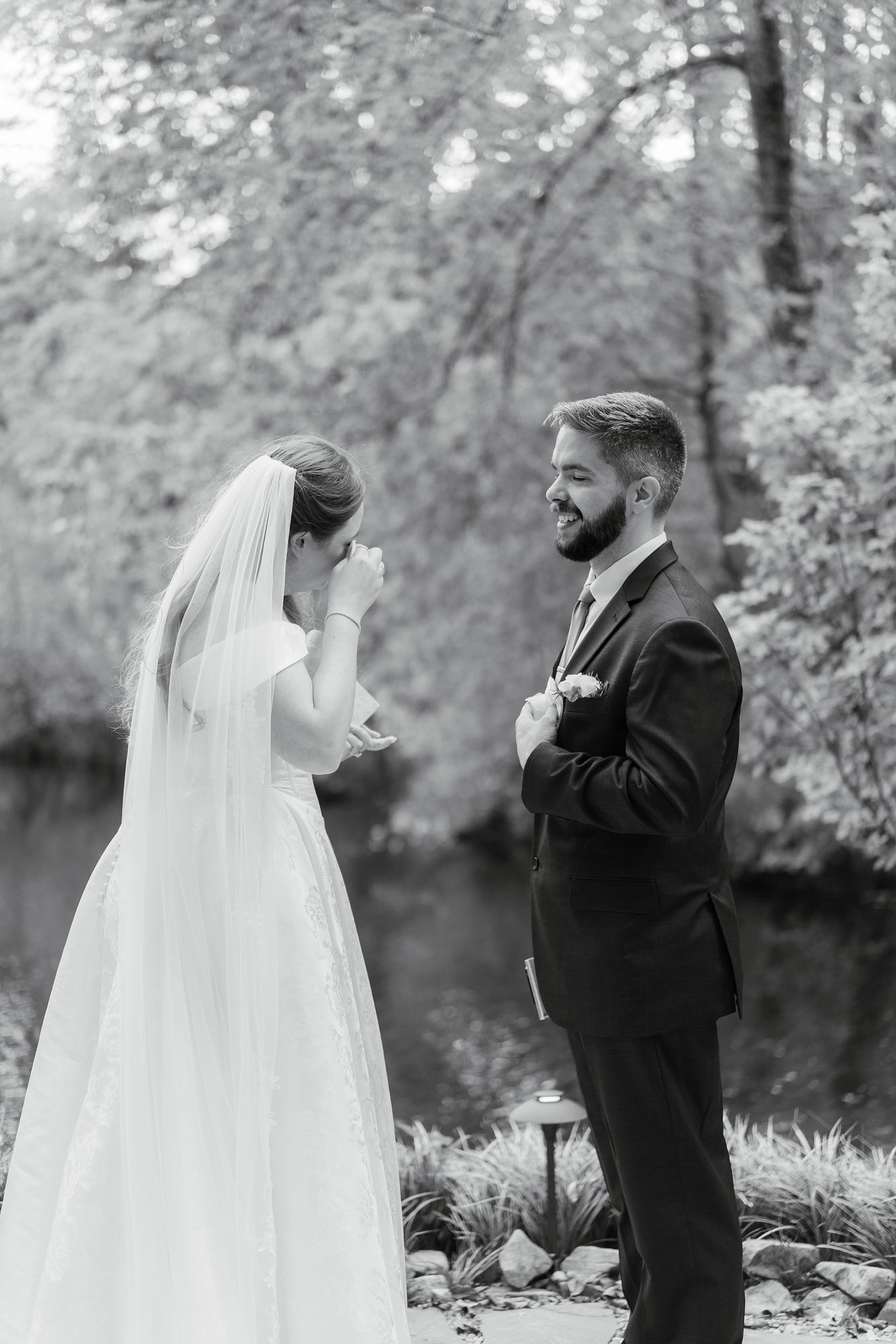 Black and white photo of emotional bride and smiling groom sharing first look moment near a tranquil pond.