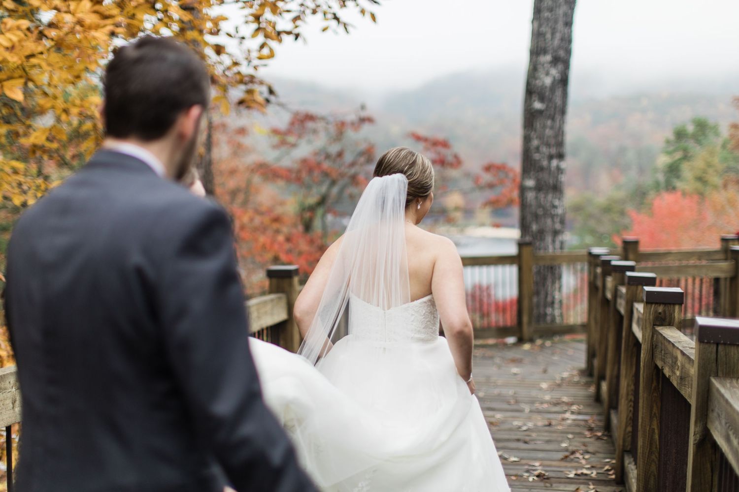 Couple in wedding attire walking along a scenic autumn mountain overlook with fall foliage in the background.