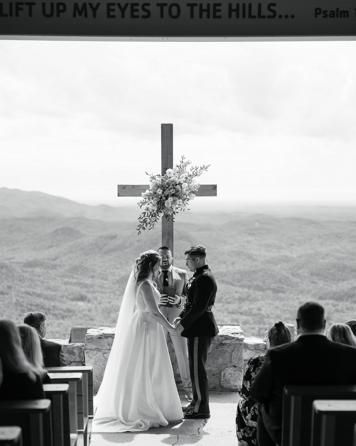 Black and white photo of an outdoor wedding ceremony atop a mountain with a large cross and guests seated on benches.