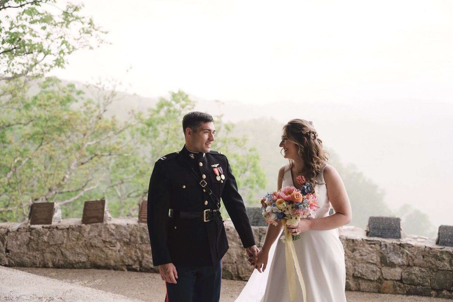 Military groom and bride share a moment while walking along a misty stone wall overlook in formal wedding attire.