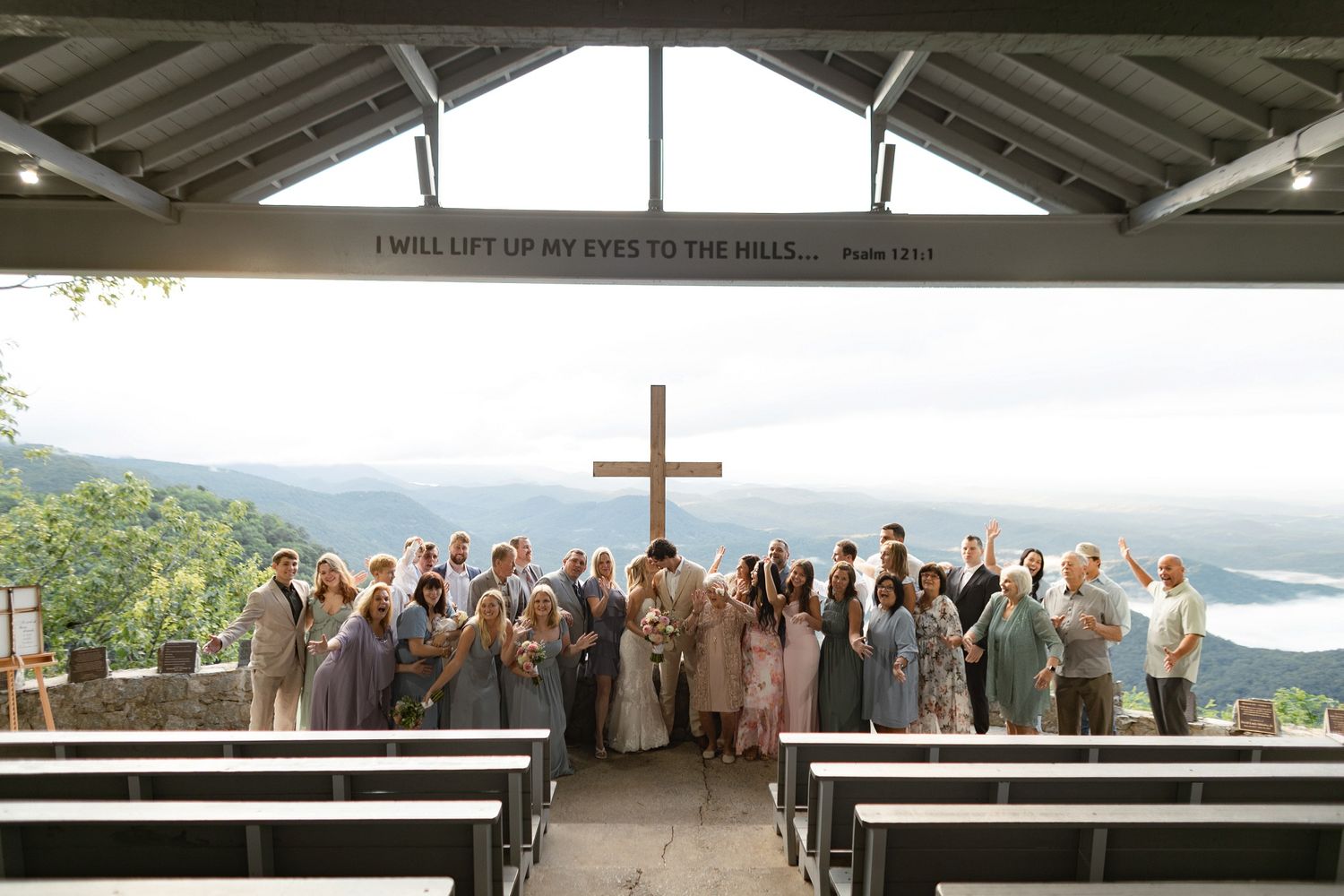 A wedding celebration group gathers at an outdoor chapel with mountain views and wooden benches under an A-frame roof.