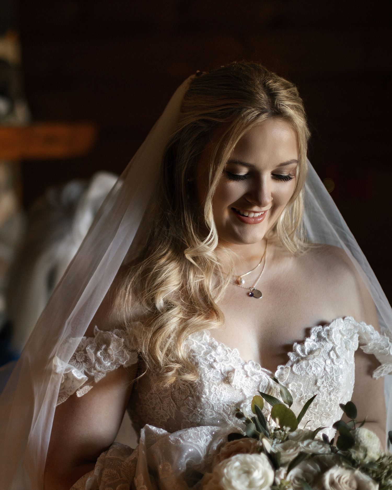 A bride in a white lace wedding dress beams with joy while holding a bouquet of roses and greenery.