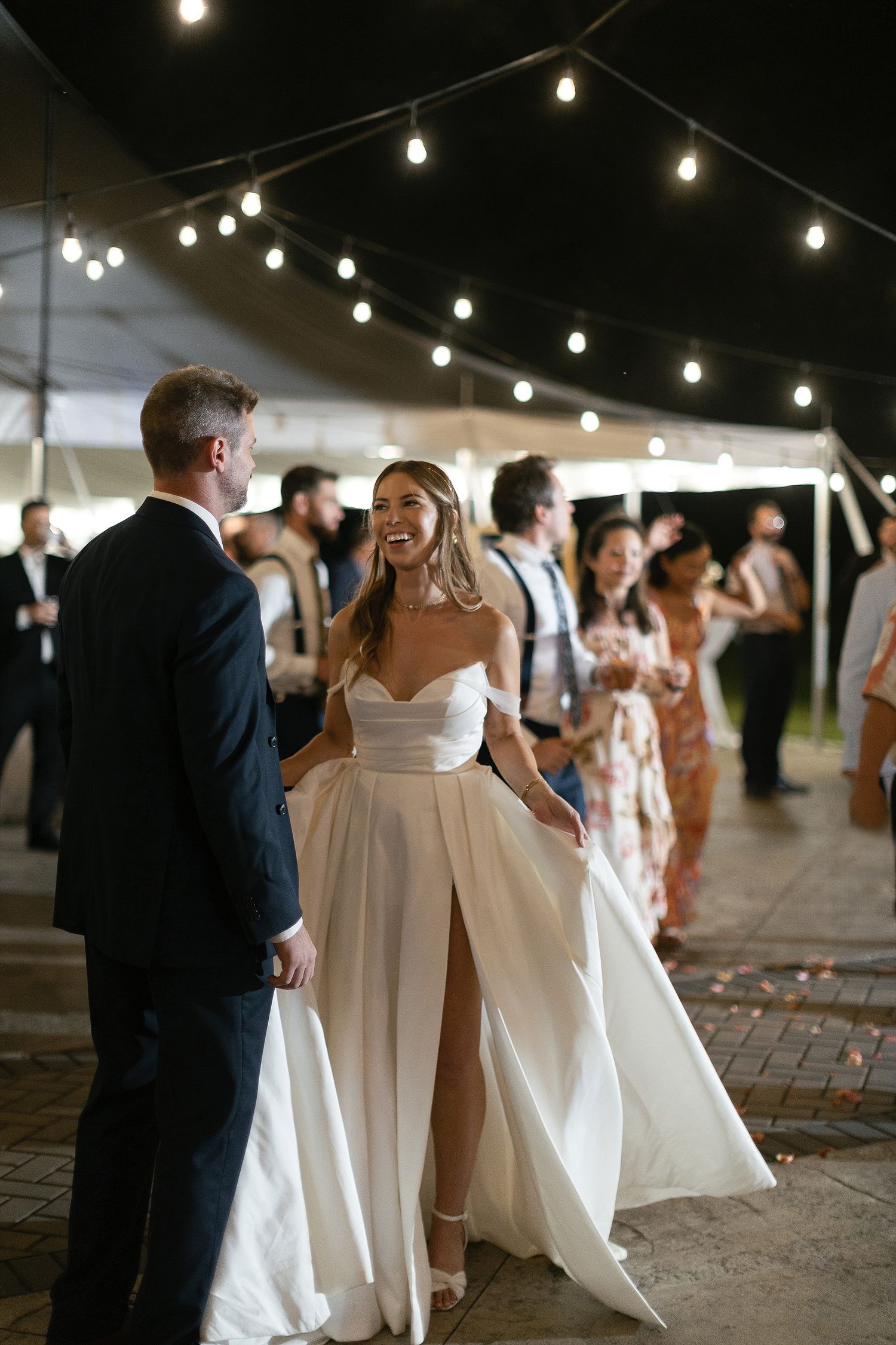 A couple dances under string lights at an outdoor wedding reception with guests in the background.