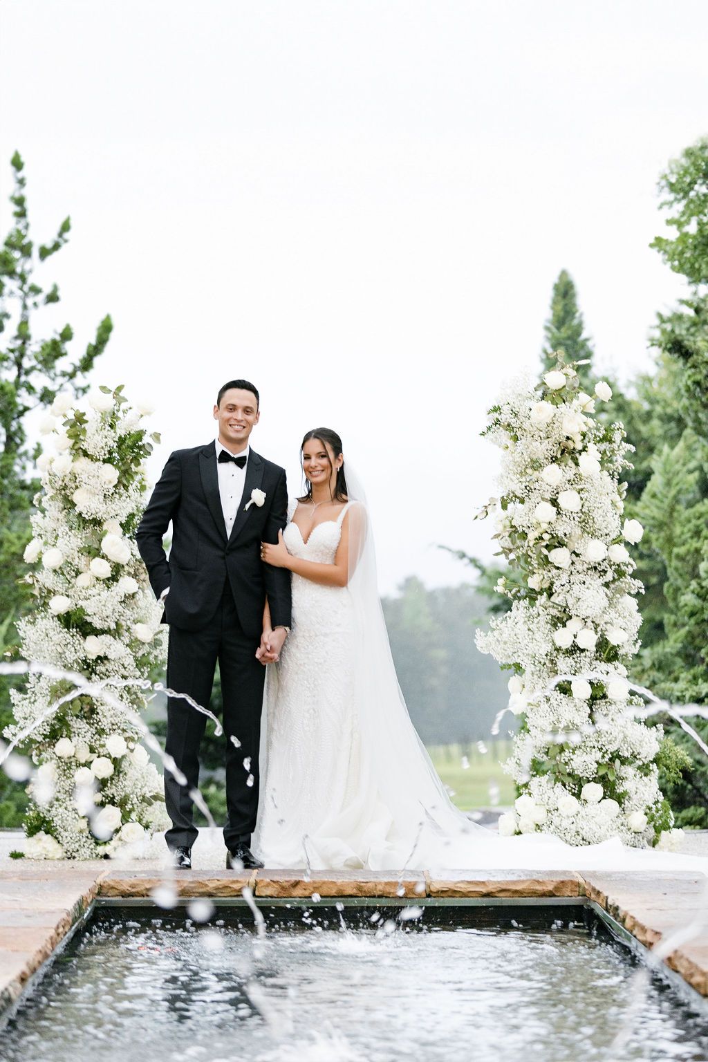 A wedding couple poses on a stone platform surrounded by white floral arrangements and fountains.