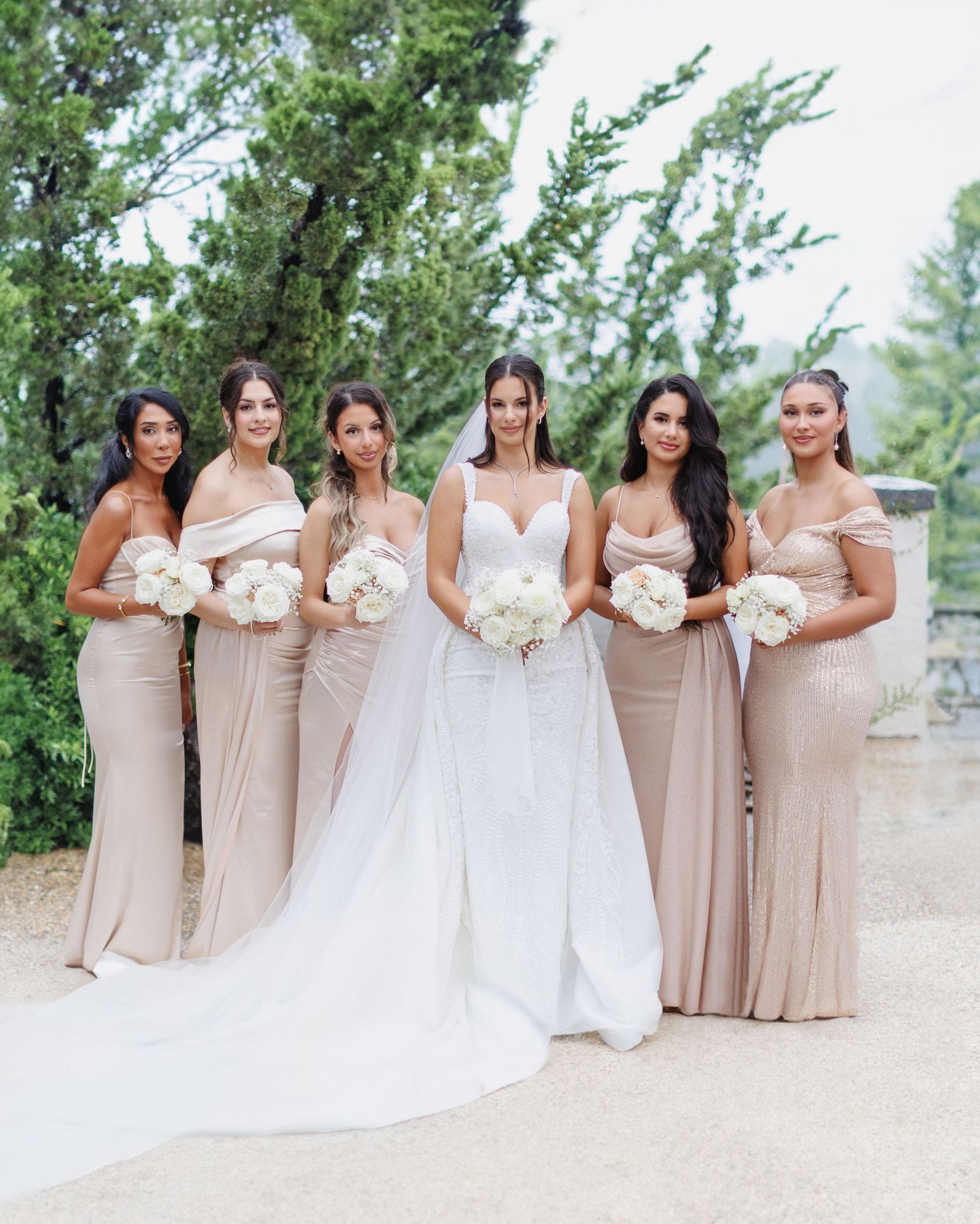 Wedding party poses outdoors in a garden setting wearing blush-colored bridesmaid dresses and white bridal gown.