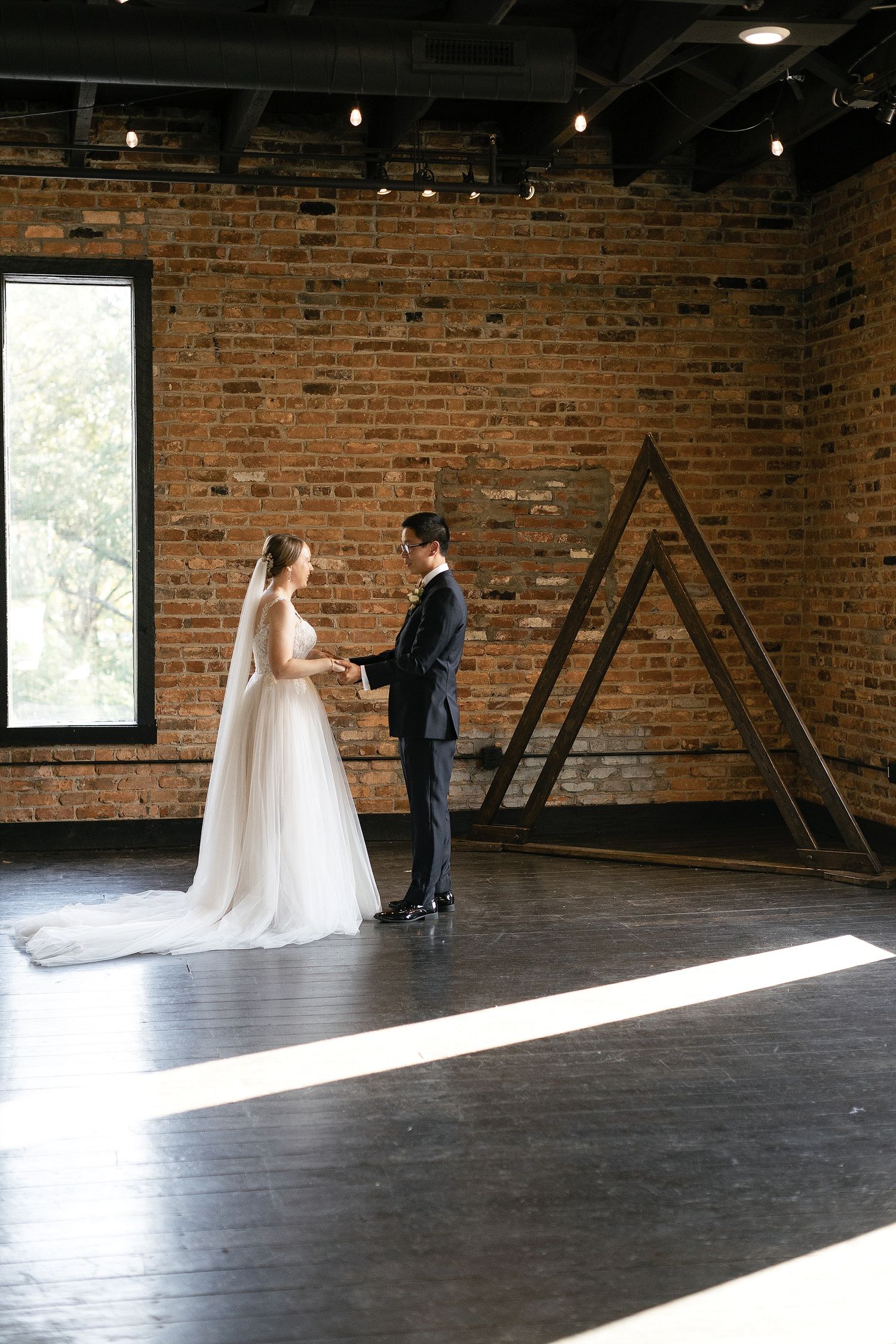 A couple holds hands in an industrial-style room with exposed brick walls, tall windows, and dramatic natural lighting.