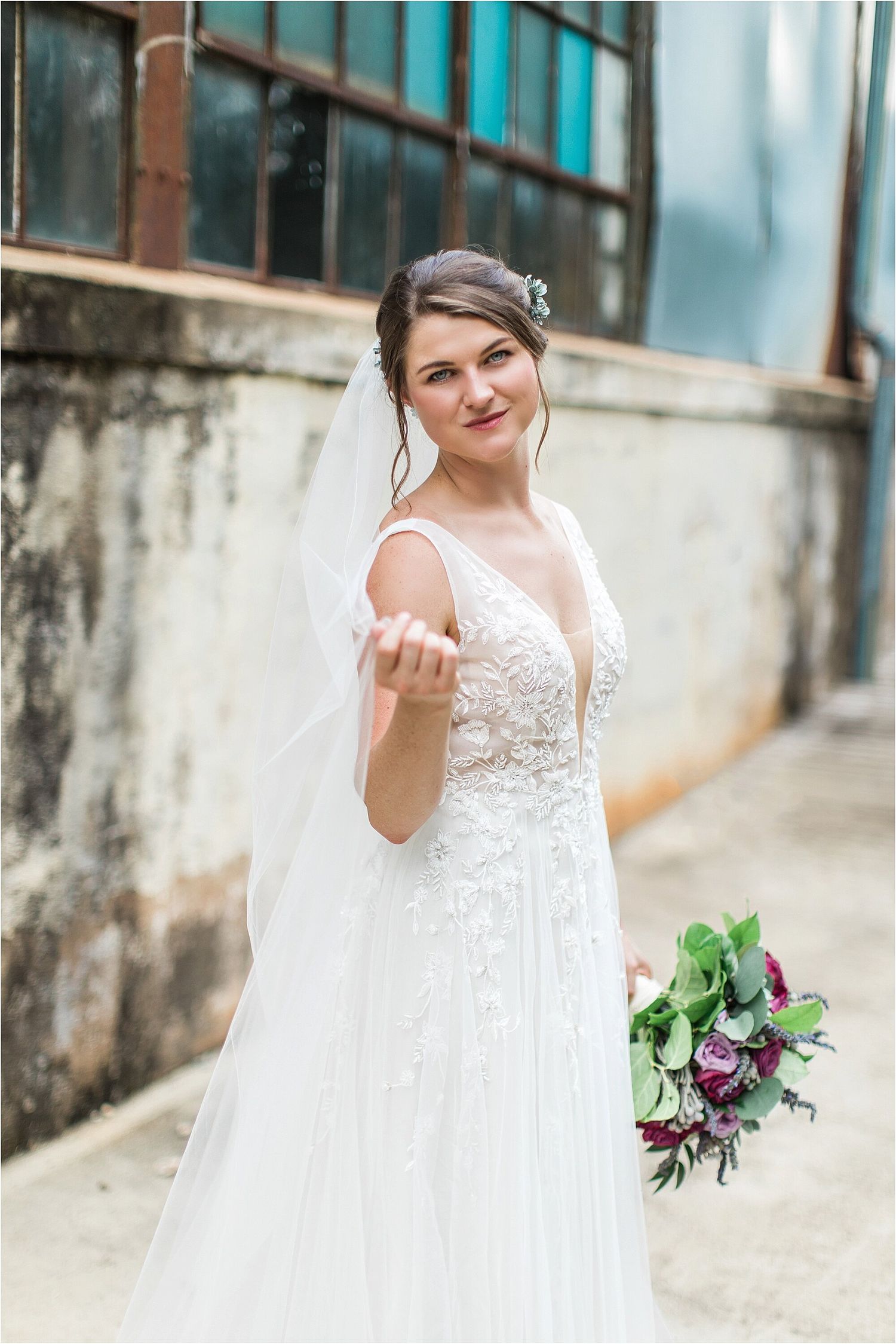 A bride in a lace wedding dress with a flowing veil holds out her hand while holding a purple and green bouquet.
