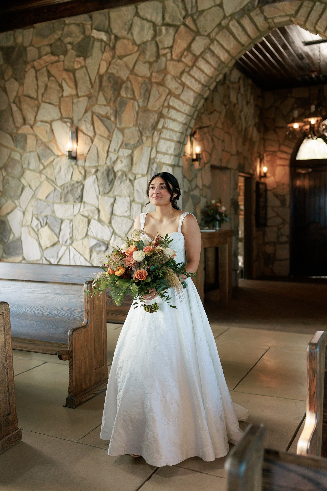 A bride in a white wedding dress sits at a table in a rustic stone-walled venue with elegant floral arrangements.