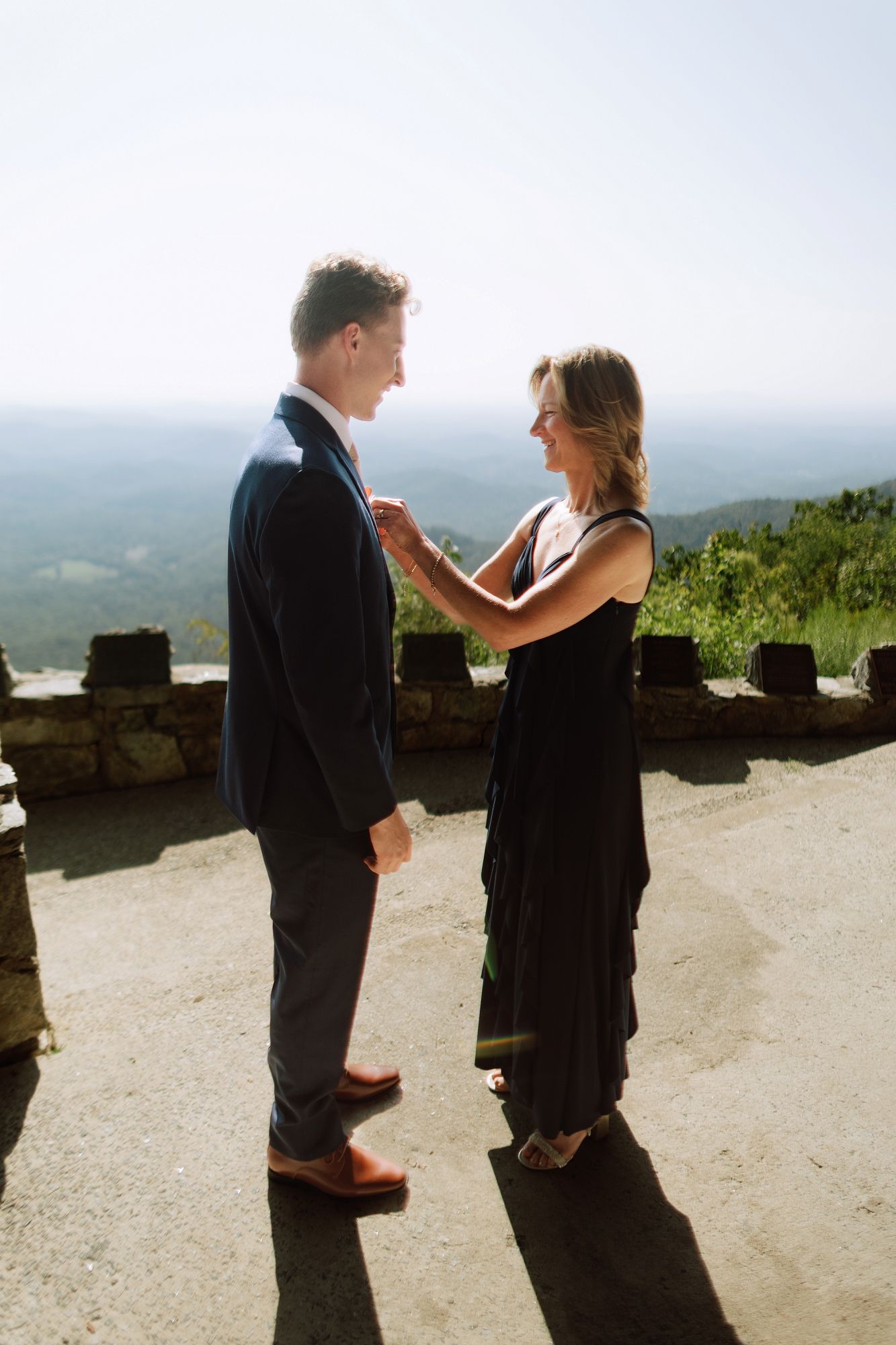 Woman in black dress adjusts grooms boutonniere on mountain overlook before wedding ceremony.