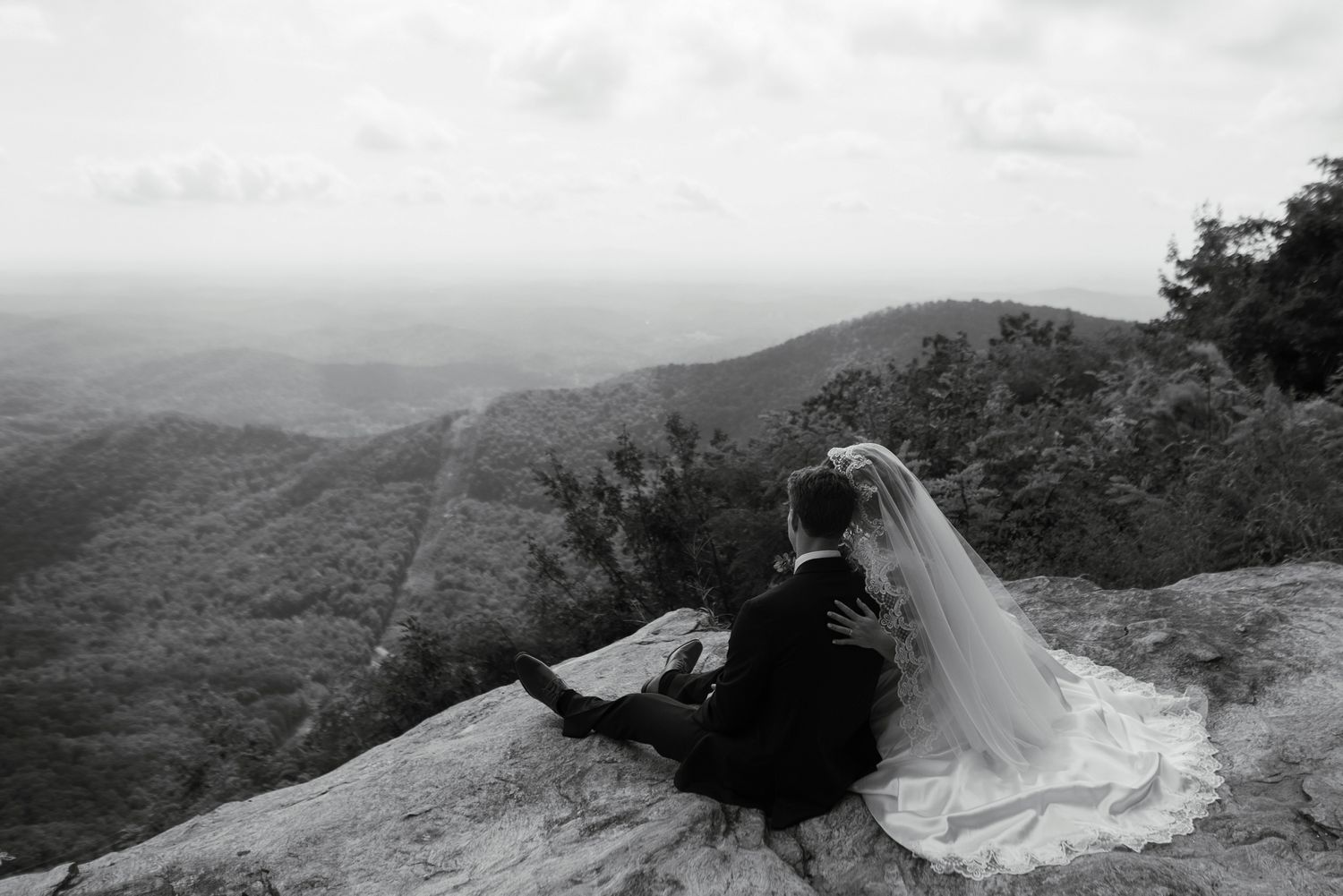 Black and white photo of bride and groom sitting on a mountain cliff edge, overlooking a vast forested valley.