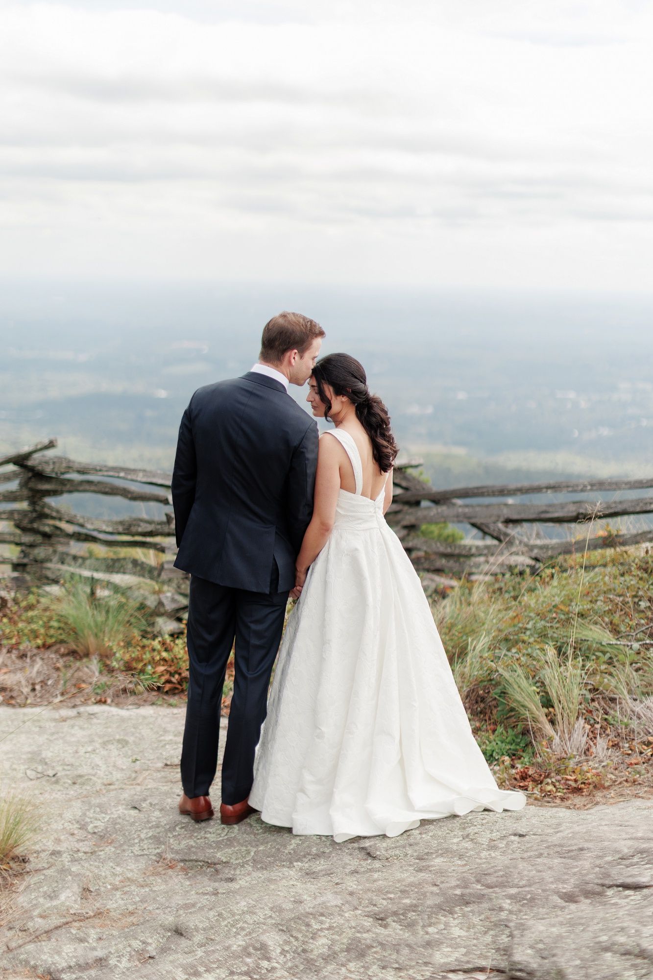 Bride and groom stand on rocky overlook with mountain valley views, she in white ballgown, he in navy suit.