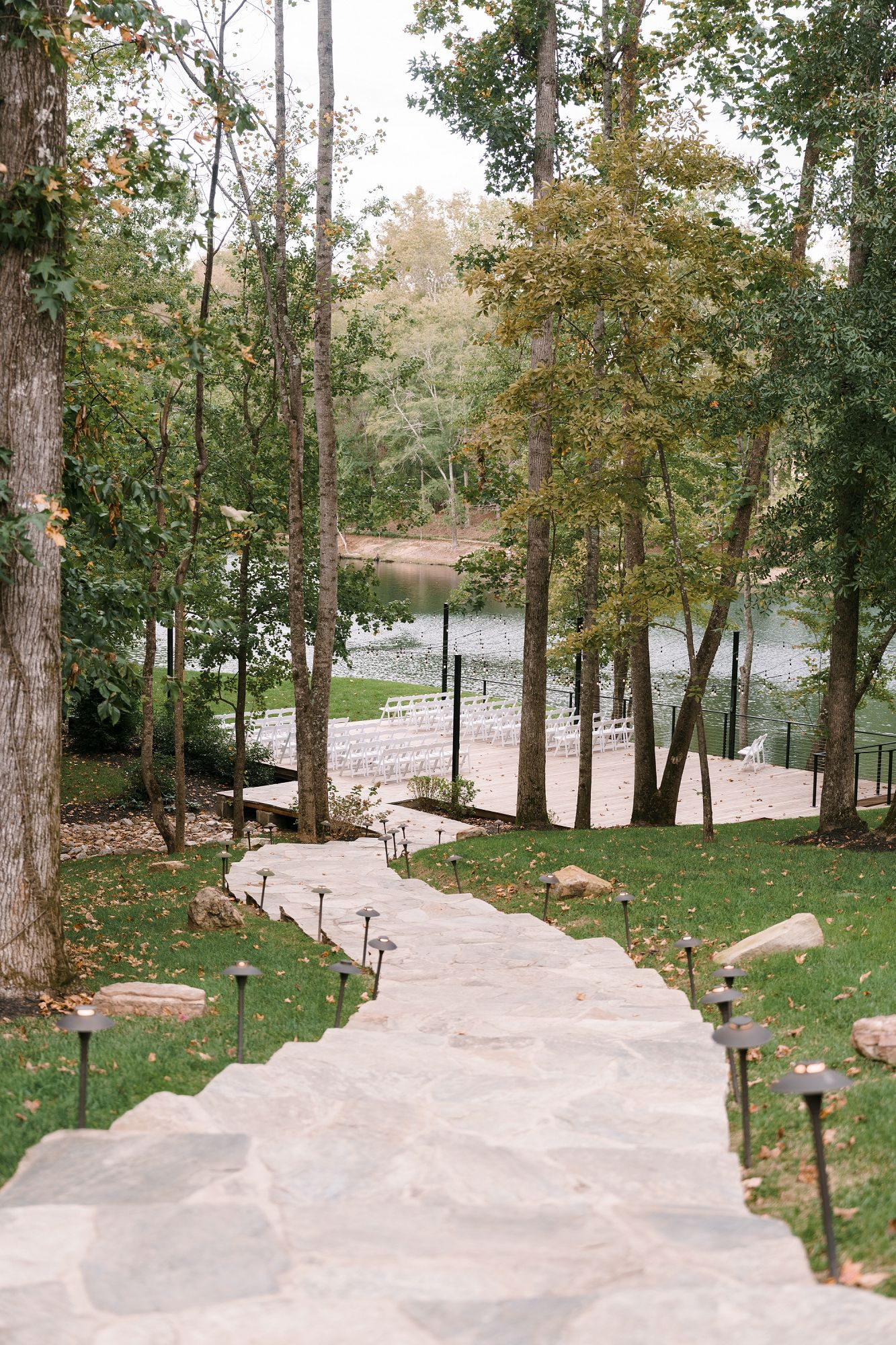 Stone pathway leads down to an elegant outdoor wedding ceremony setup beside a serene lake surrounded by lush trees.