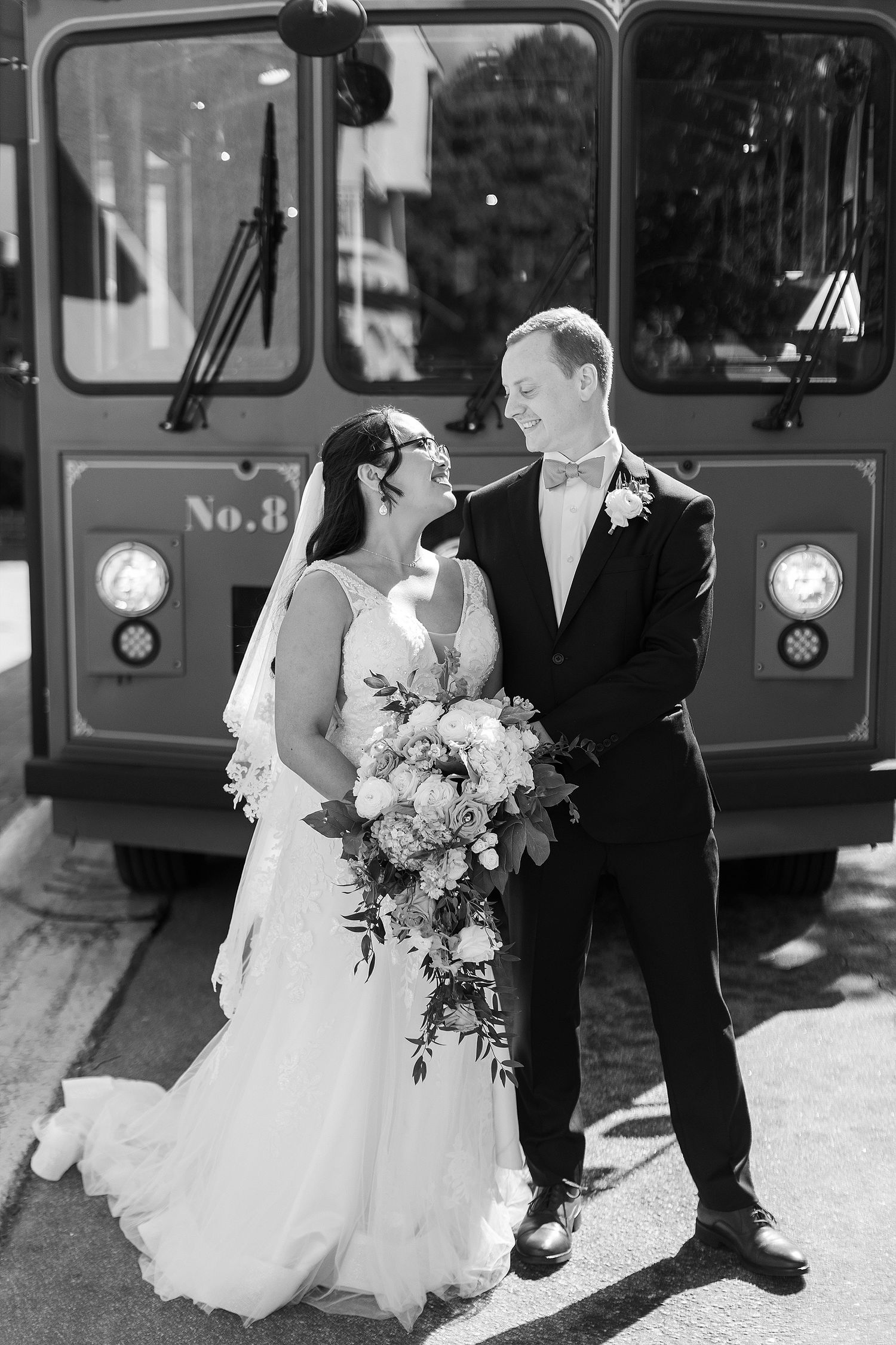 A classic black and white wedding photo of a couple posing in front of a vintage trolley.