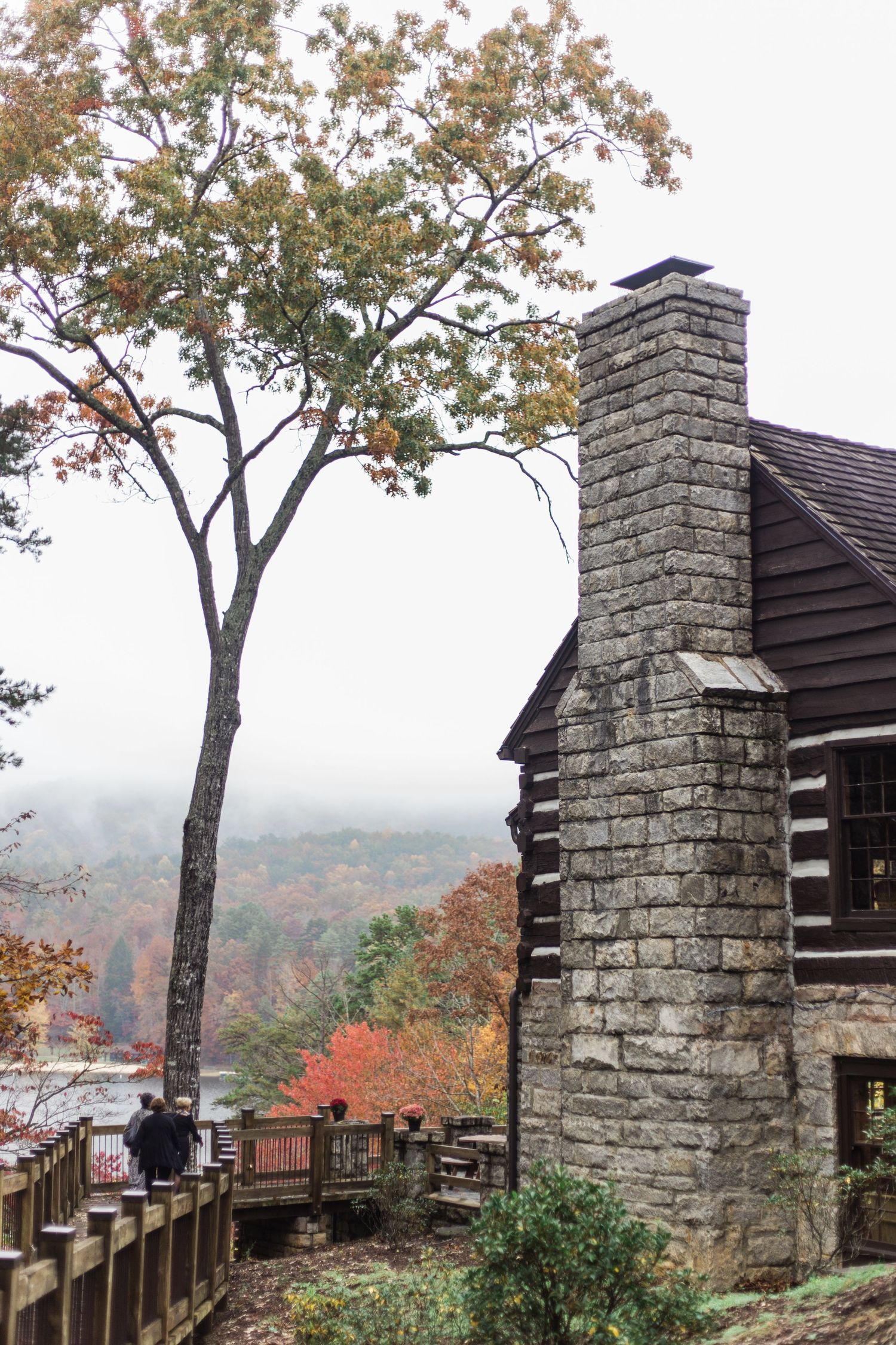 A rustic log cabin with stone chimney overlooks a misty autumn valley from a wooden deck surrounded by fall foliage.