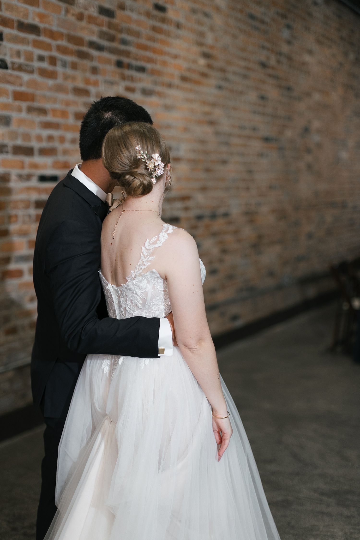 Beautiful wedding portrait shows couple embracing against brick wall with bride in white lace gown with elegant updo hairstyle.
