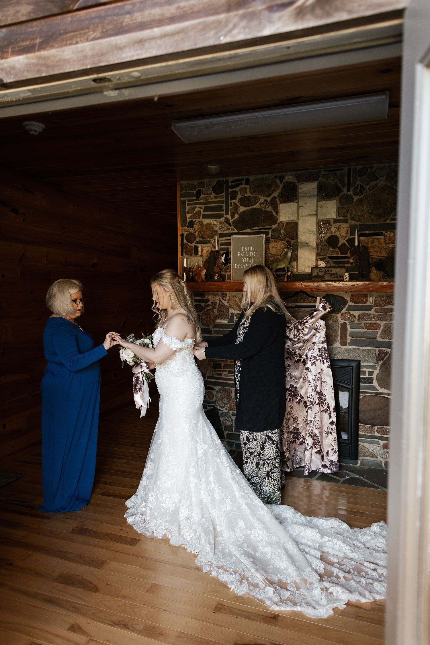 A bride wearing a flowing lace wedding gown with long train gets ready in a rustic stone-walled room with hardwood floors.