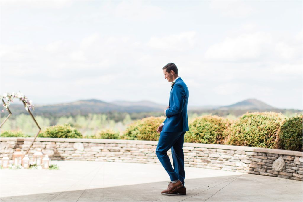 Groom in blue suit stands contemplatively on stone patio overlooking mountain vista.