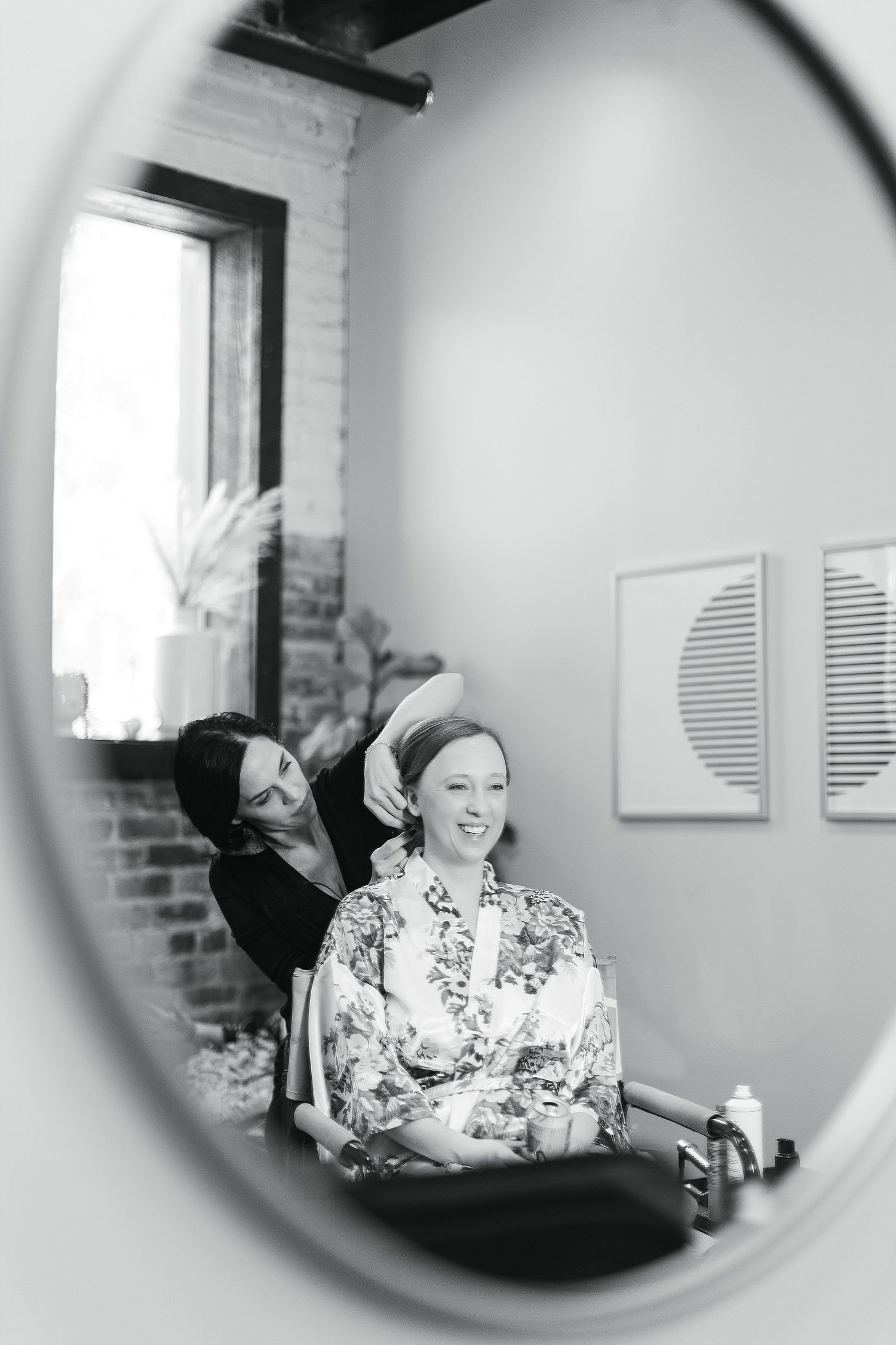 Black and white reflection in mirror shows someone getting their hair styled while wearing a floral robe during preparation.