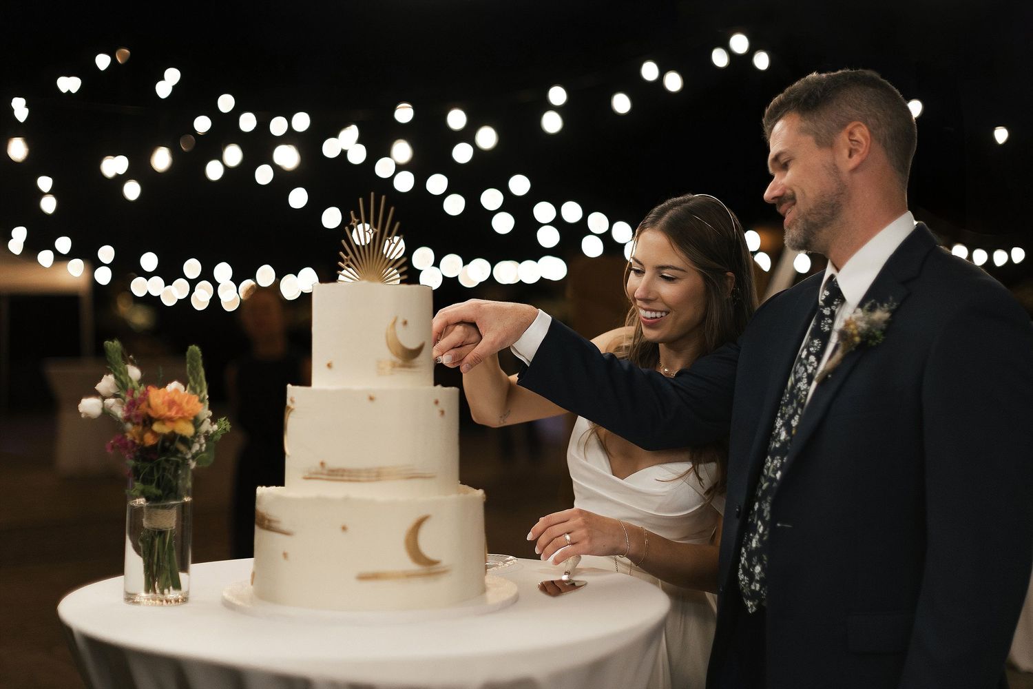 Couple cuts elegant celestial-themed wedding cake under twinkling lights.
