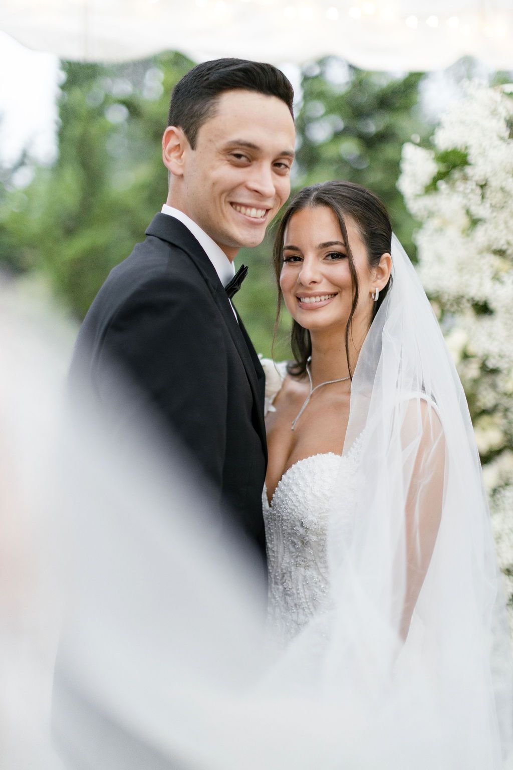 A joyful wedding portrait captured outdoors with white floral decorations in the background.