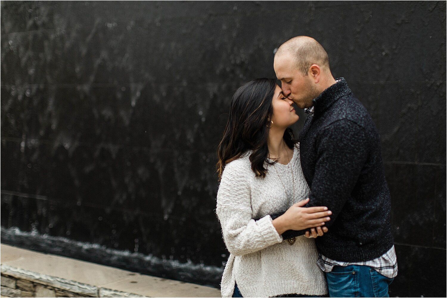 Intimate winter engagement photo of couple embracing against dark wall in snowfall.