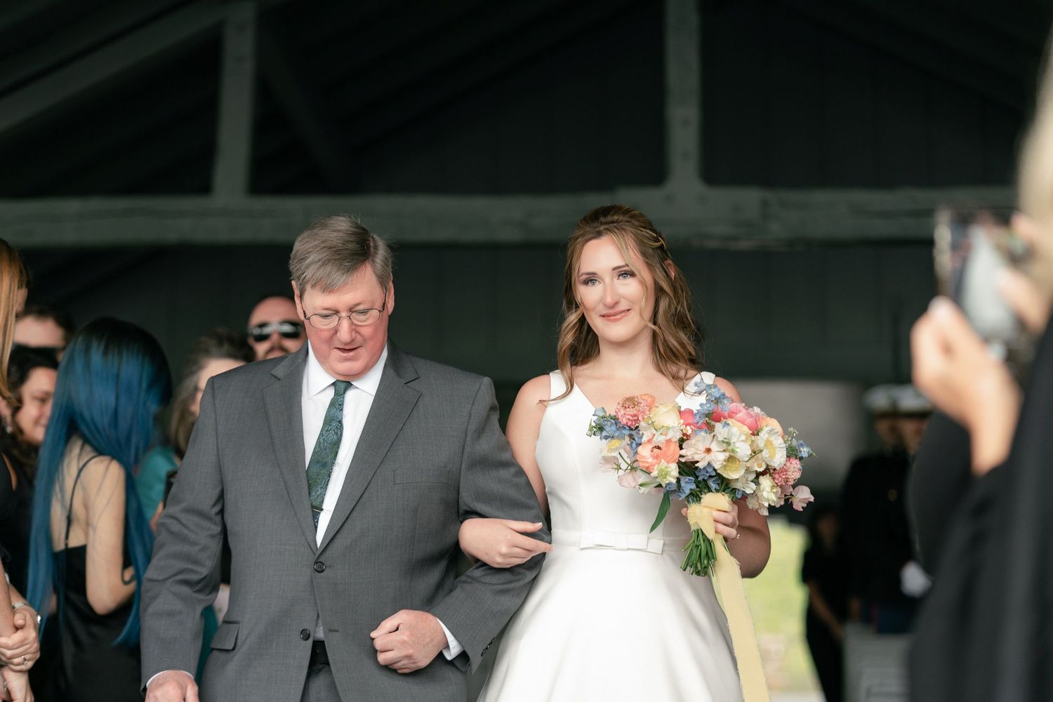 A couple walking down the aisle at an outdoor wedding ceremony, the bride wearing a white dress and holding a bouquet.
