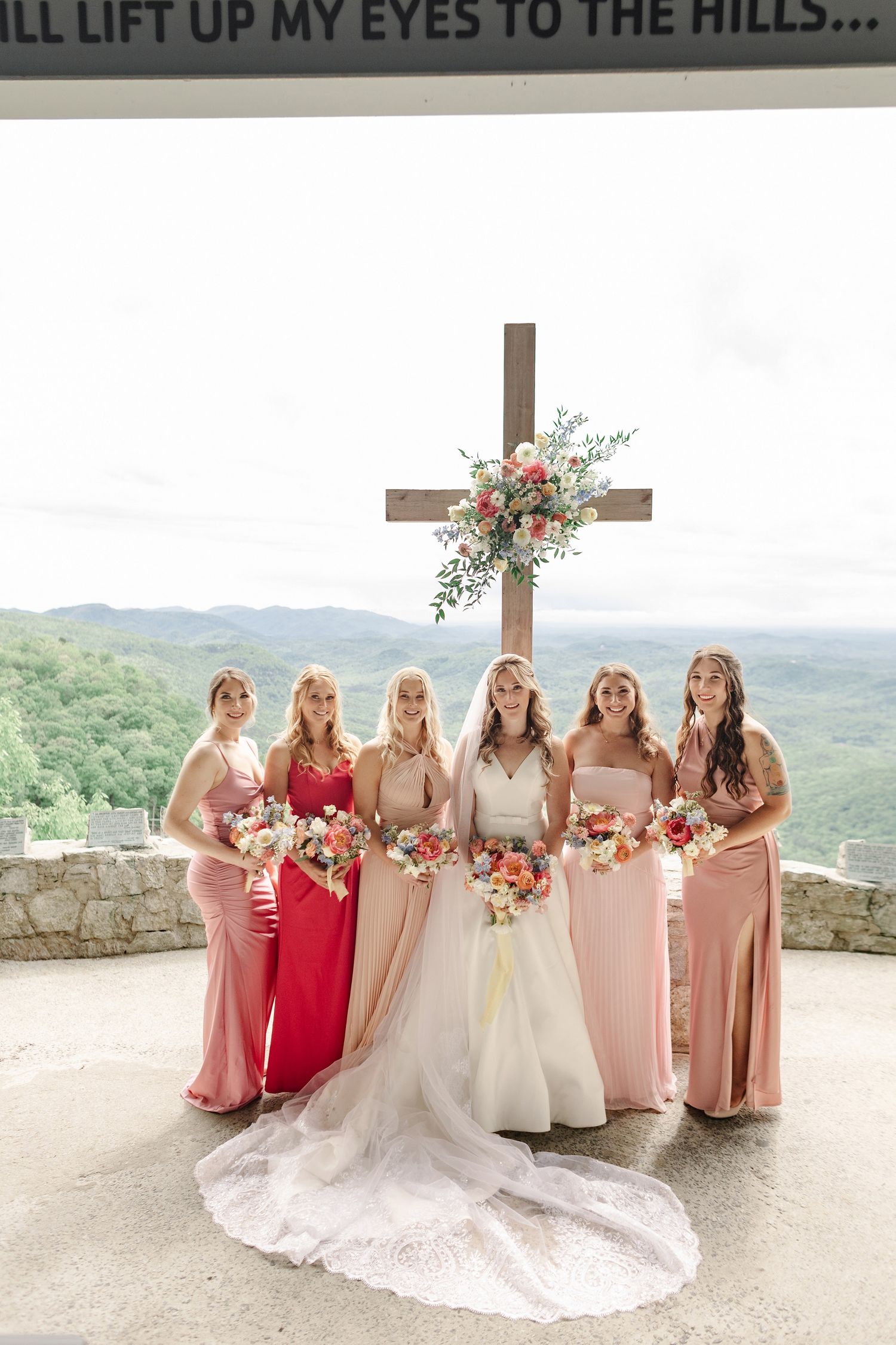 A wedding party in pink and red dresses poses against a scenic mountain backdrop at a stone overlook point.