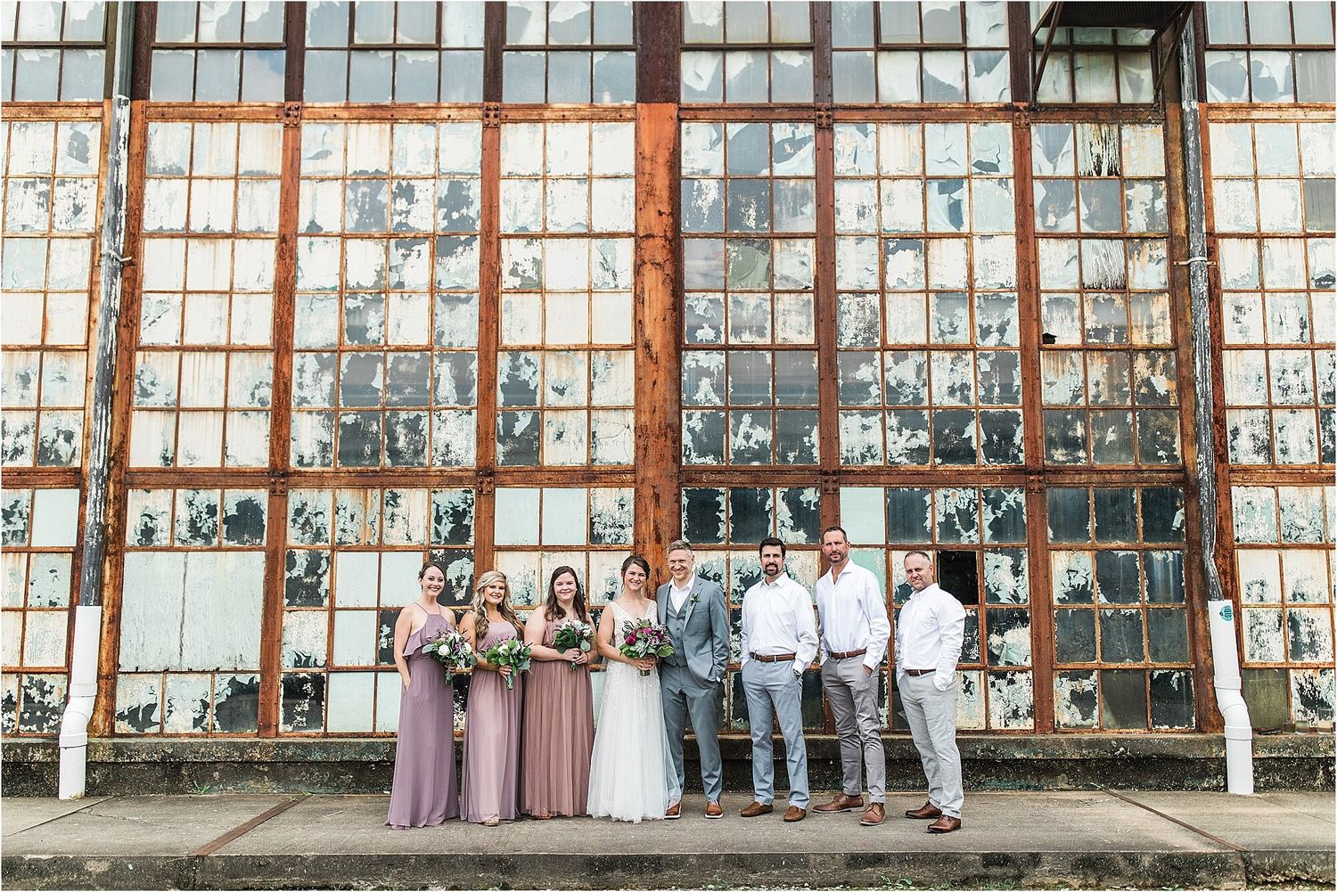 Wedding party poses in front of large industrial windows wearing purple and grey attire.