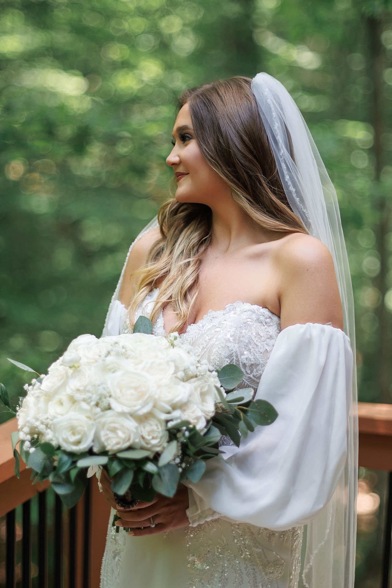 Bride holding white rose bouquet poses in off-shoulder gown on outdoor deck.