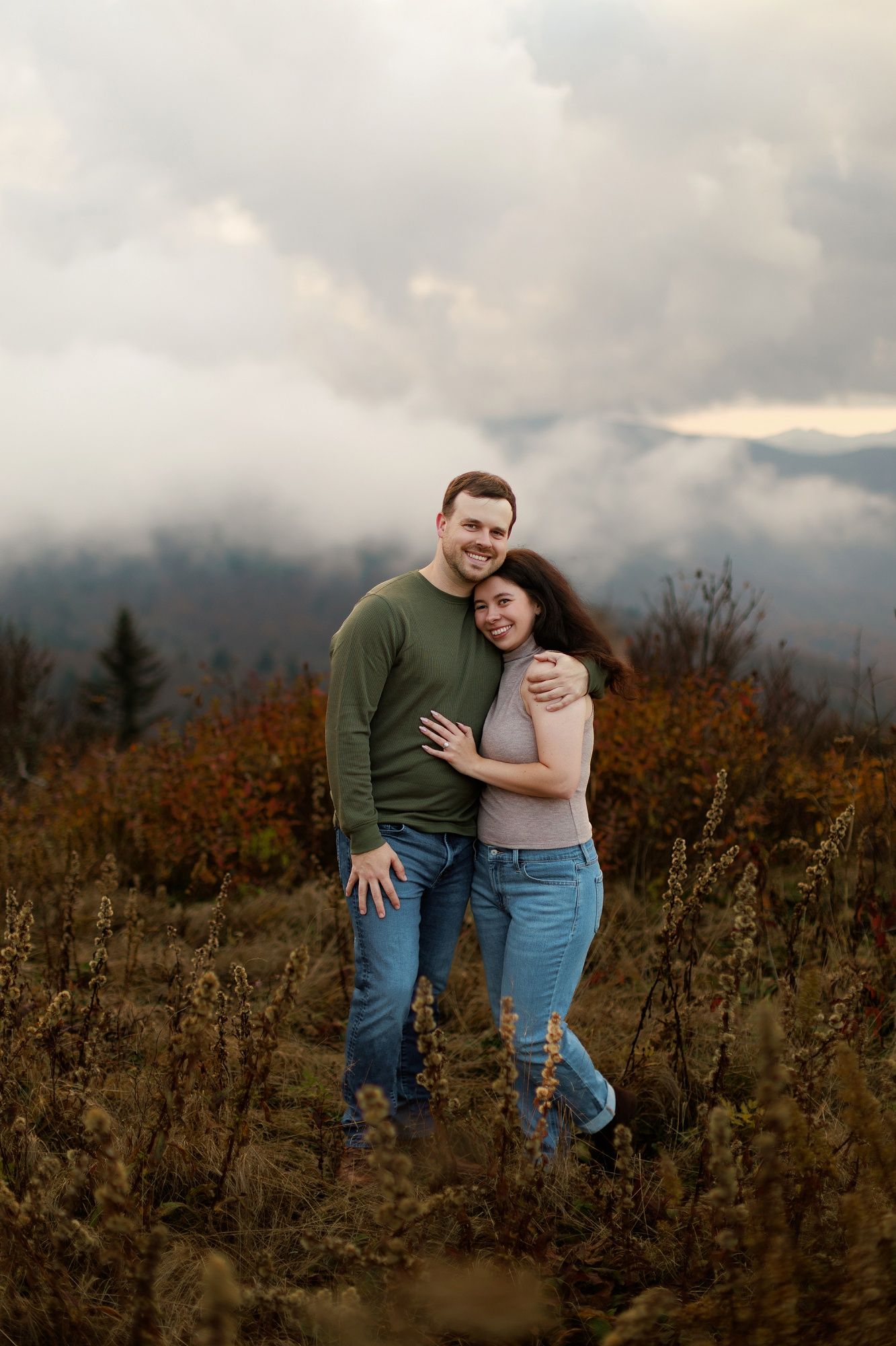 A couple embraces on a misty mountainside with autumn foliage and cloudy skies in the background.