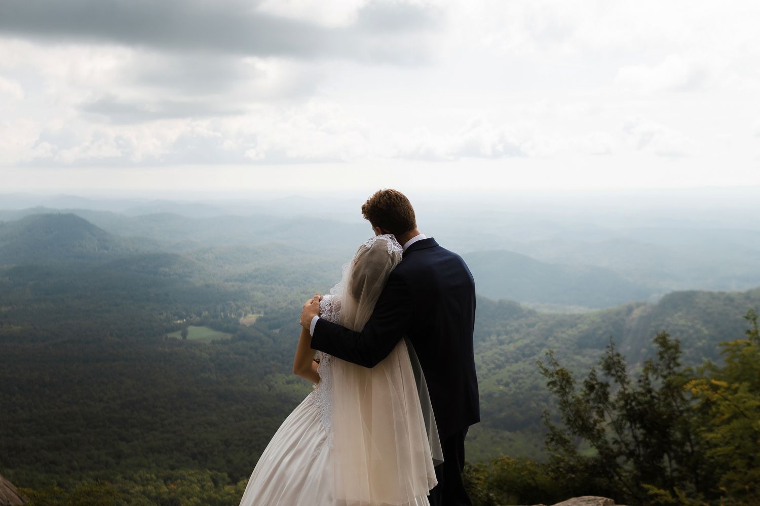 Bride and groom embrace while overlooking stunning green mountain landscape on their wedding day.