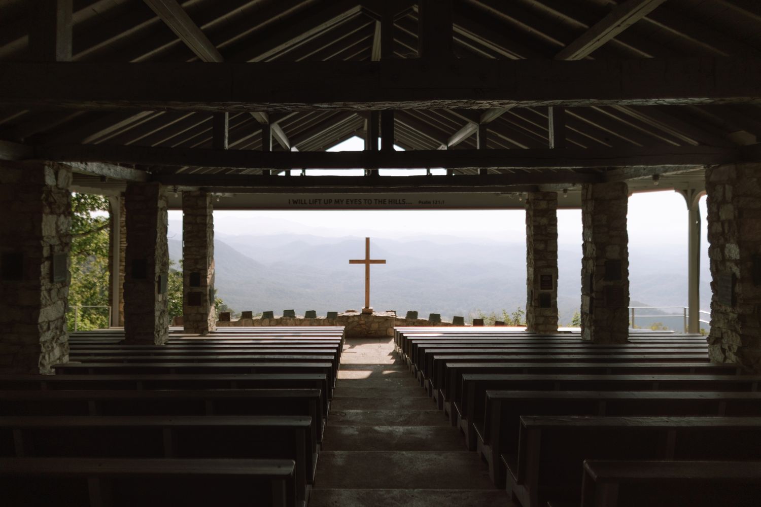 Empty open-air stone chapel with wooden pews and a cross overlooking misty mountain landscape.