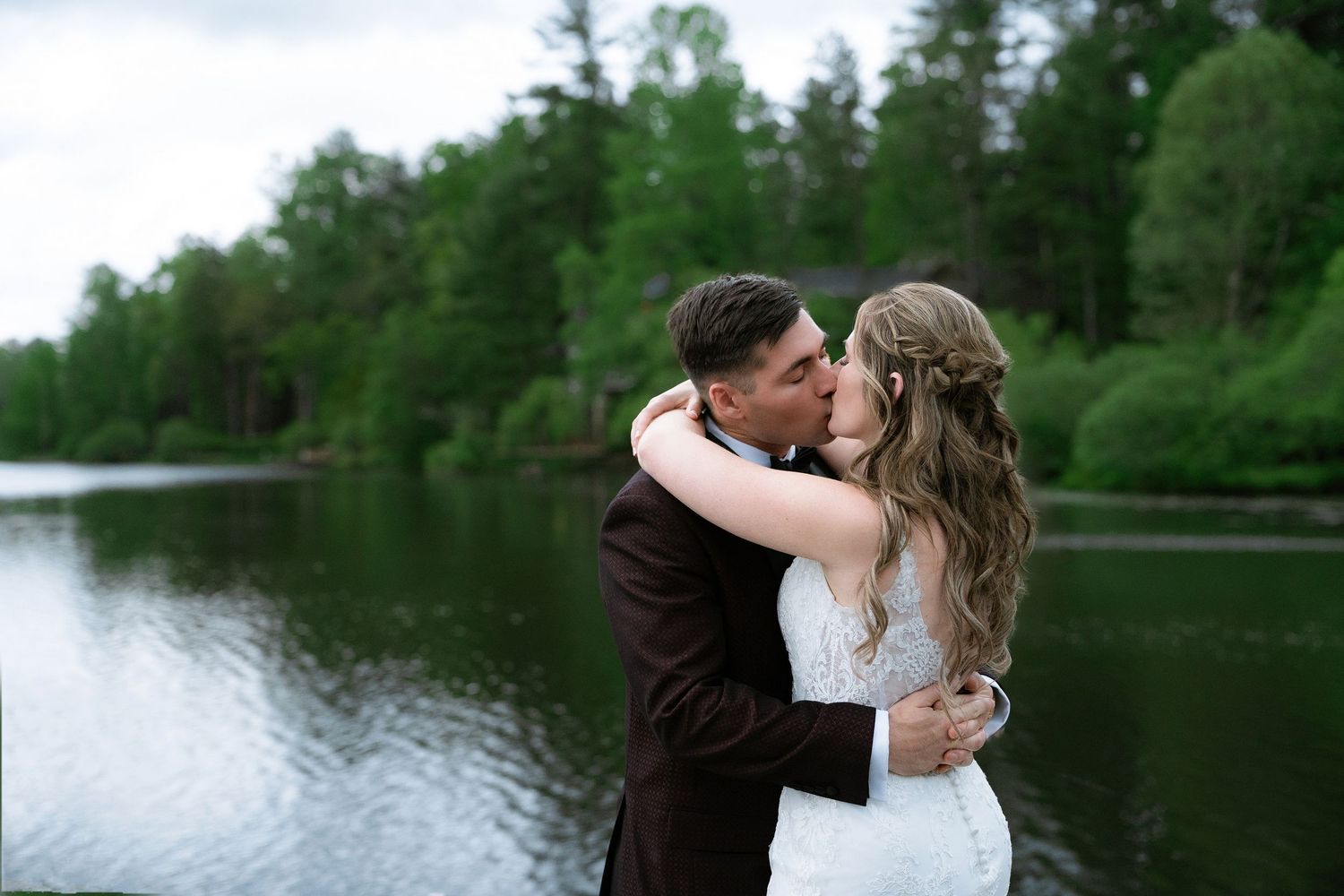 A couple shares a romantic kiss by a serene lake surrounded by green trees on their wedding day.