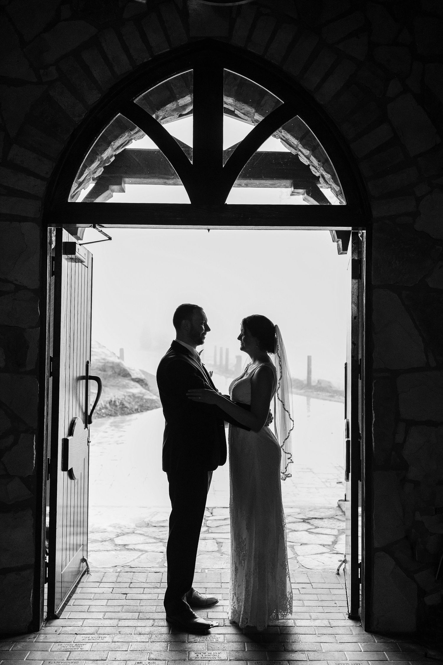 Silhouetted couple standing in arched doorway of stone chapel.