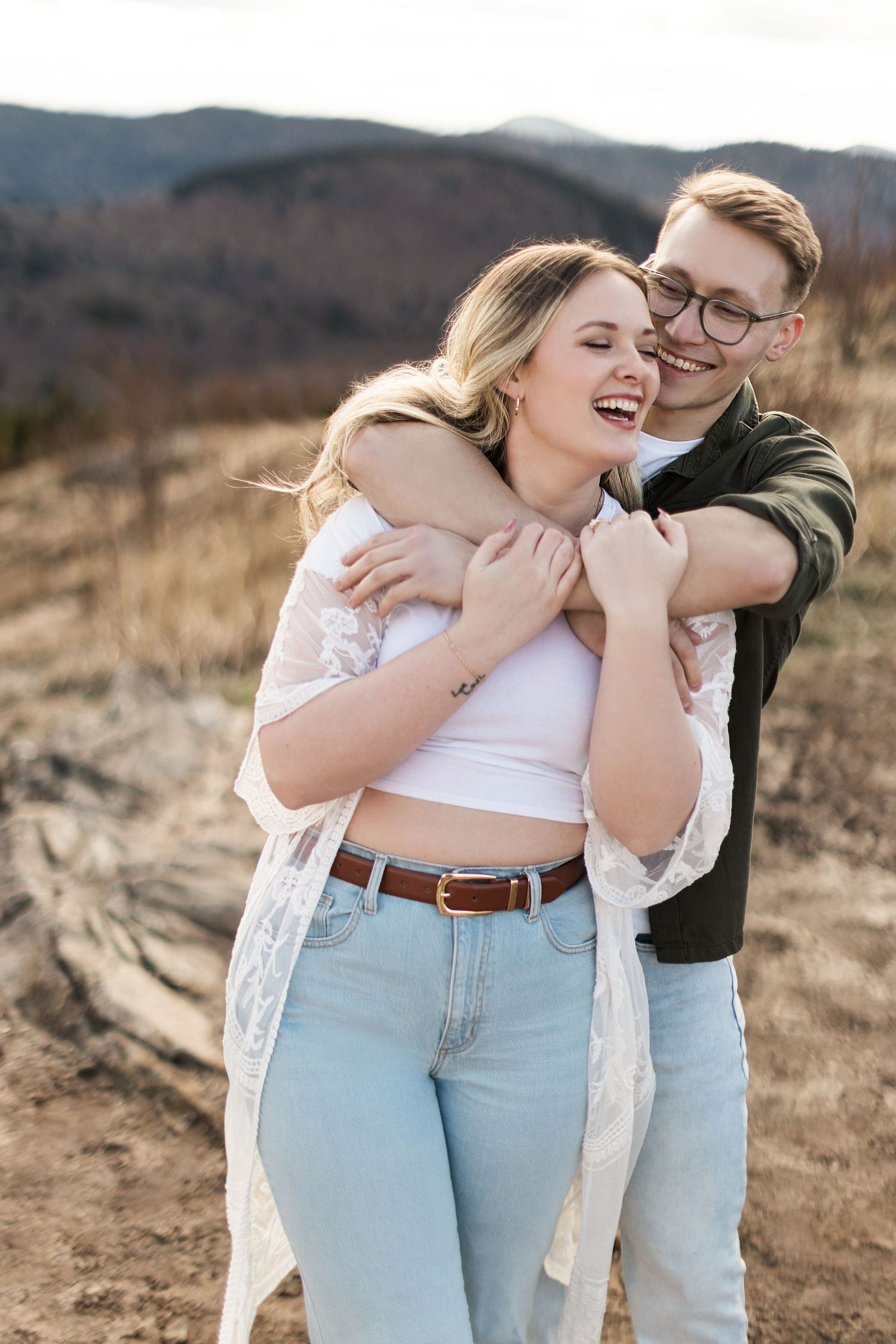 A couple embraces and laughs together while standing in a desert landscape with mountains in the background.