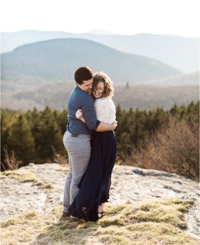 Couple embracing on mountain summit with rolling hills in background.