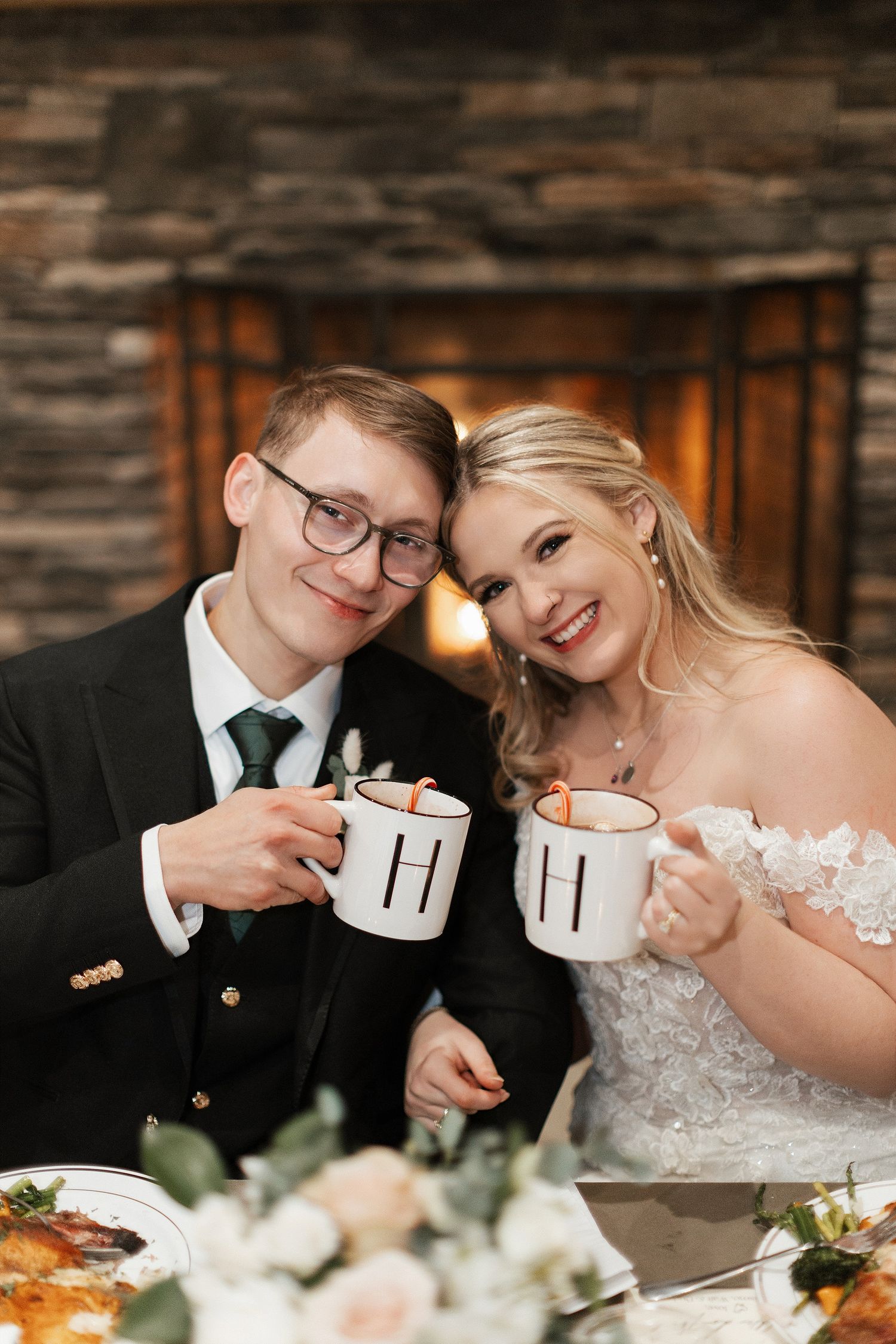 Newlywed couple toasts with matching monogrammed mugs at rustic reception with floral decor.