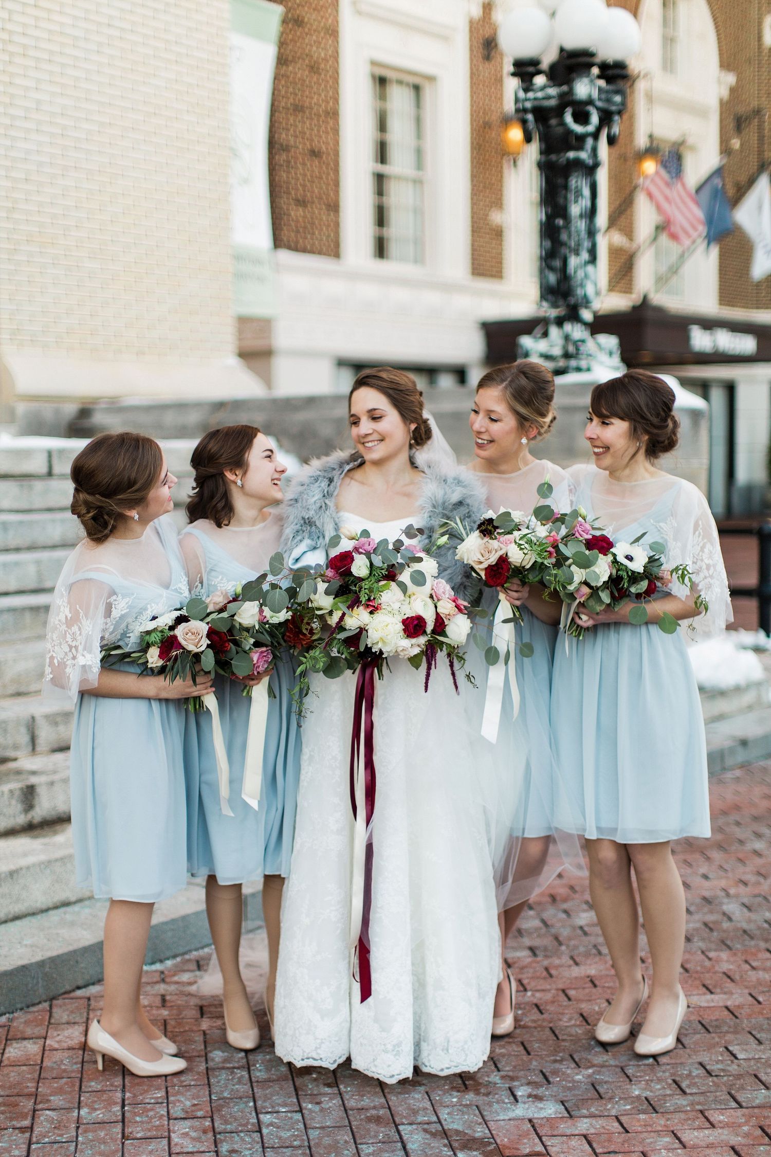 Bridesmaids in light blue dresses holding burgundy and white floral bouquets on brick pavement.