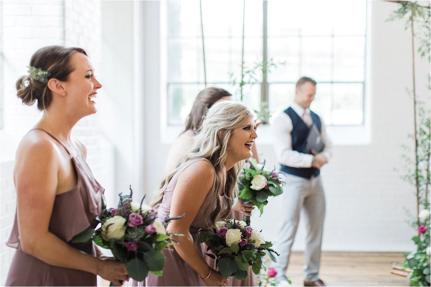 Bridesmaids in mauve dresses hold floral bouquets while laughing in a bright indoor venue.